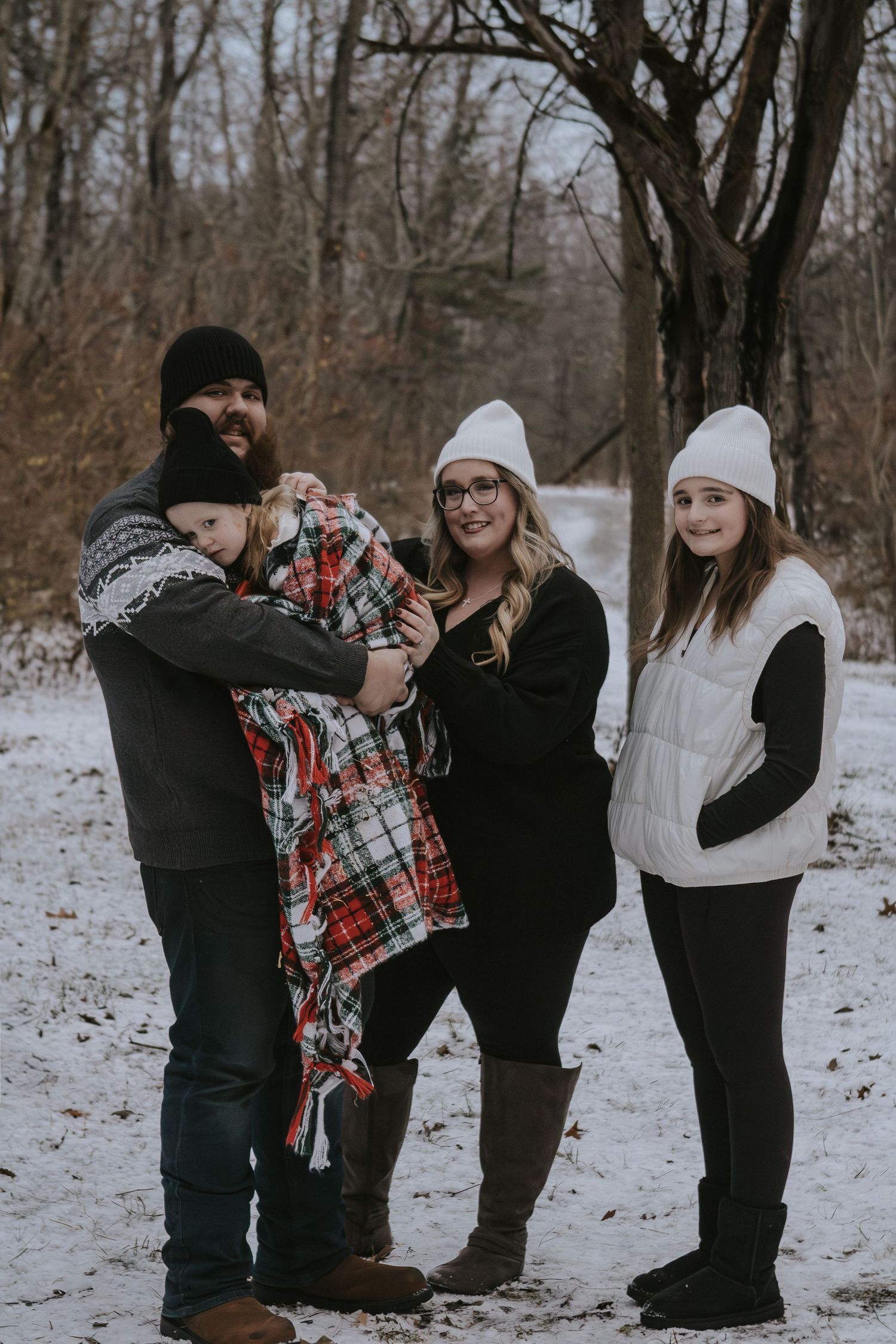 A family of four poses in a snowy winter setting, wearing cozy hats and warm clothing among bare trees.