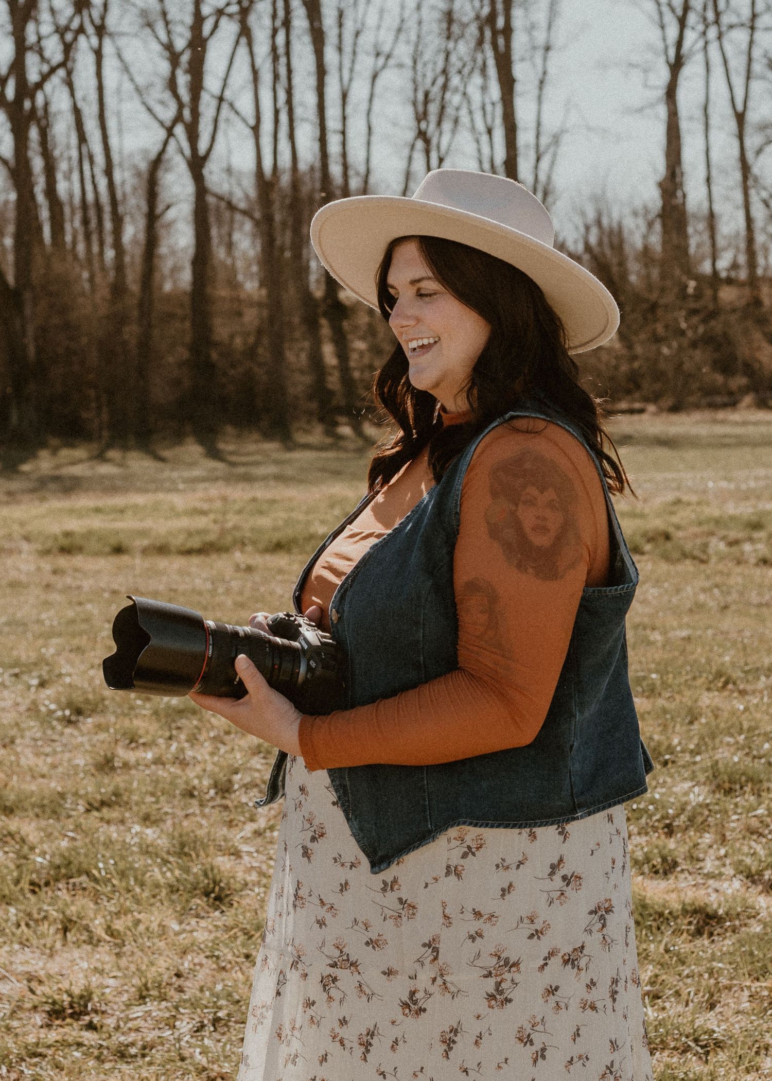 Female photographer smiling outdoors holding camera, wearing hat and denim vest in golden light.