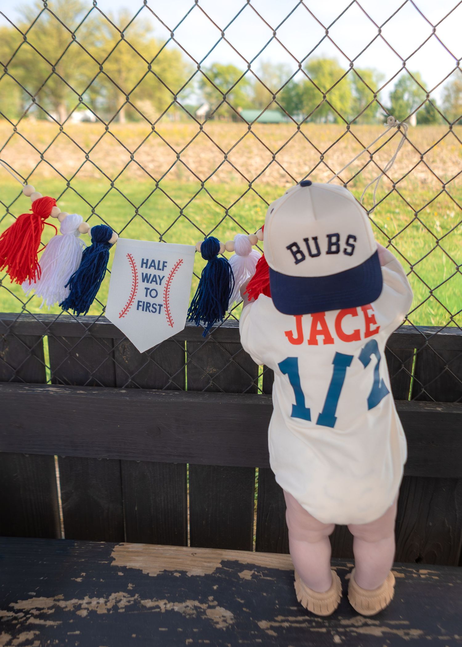 Baby in personalized Jace baseball outfit and Bubs cap at a baseball field fence.