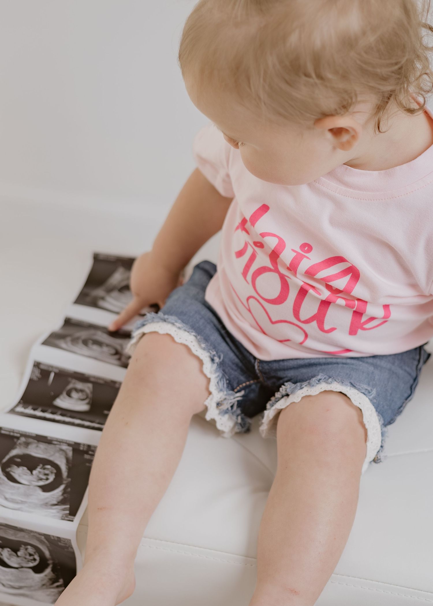 Toddler girl in pink Big Sister shirt sitting on white surface looking at ultrasound photos.