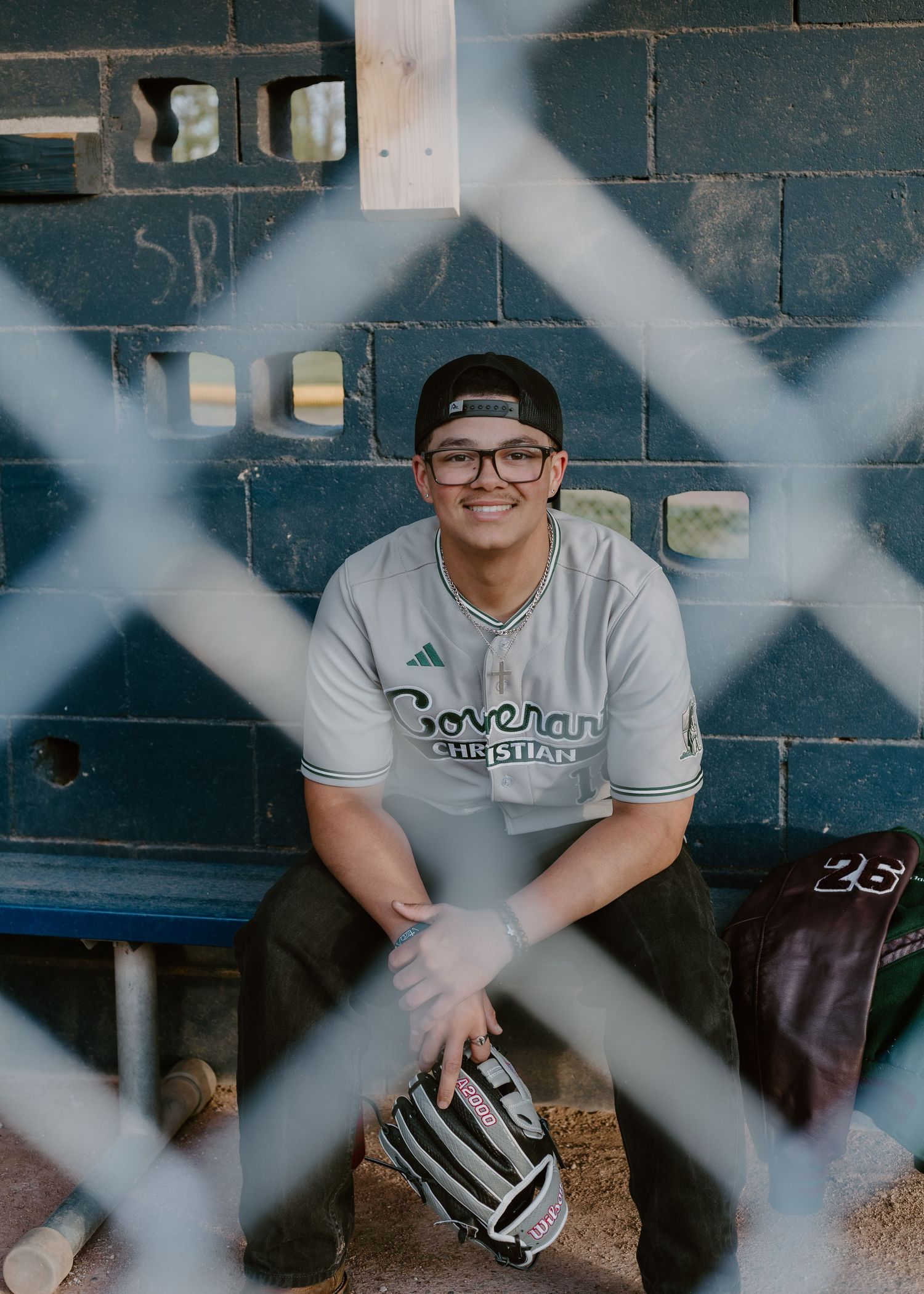 Smiling baseball player in Covenant Christian jersey sitting in dugout with glove, photographed through chain-link fence.