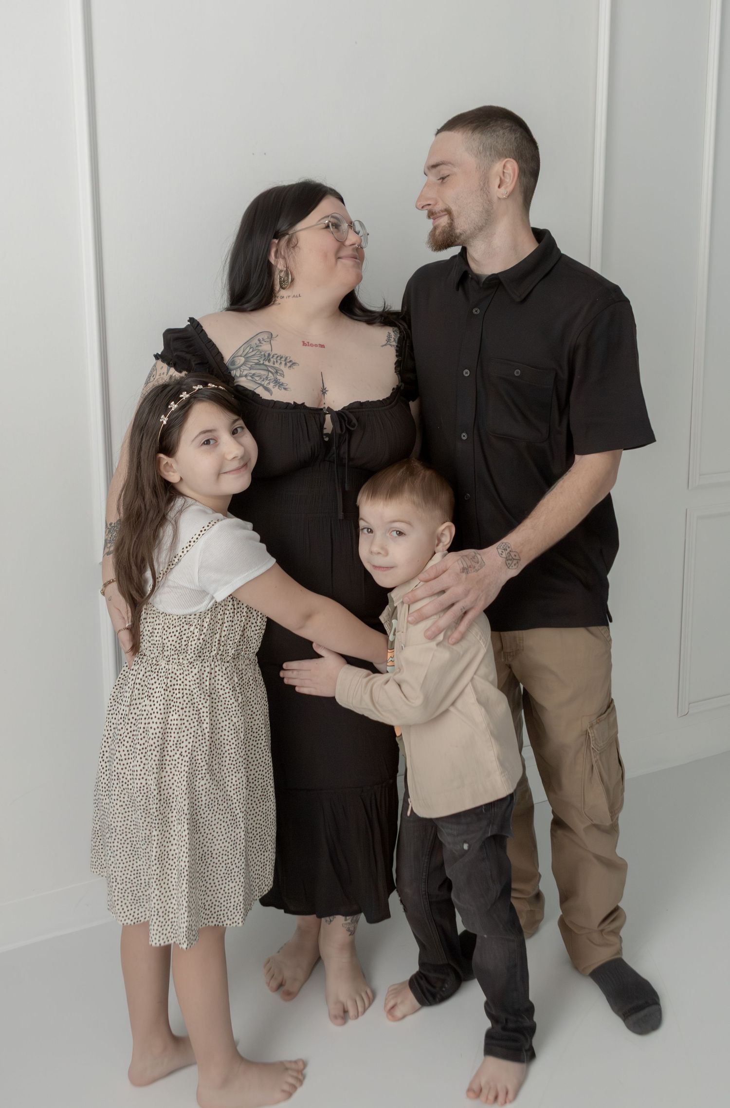 Family of four posing together in a white studio, expecting mother in a black maternity dress.
