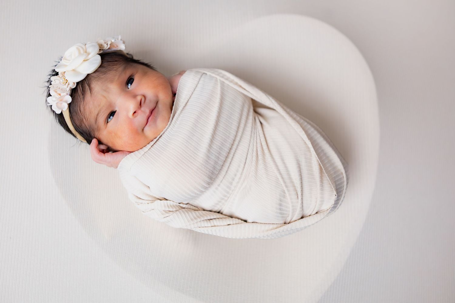 Smiling newborn baby swaddled in cream wrap with floral headband in white bowl.