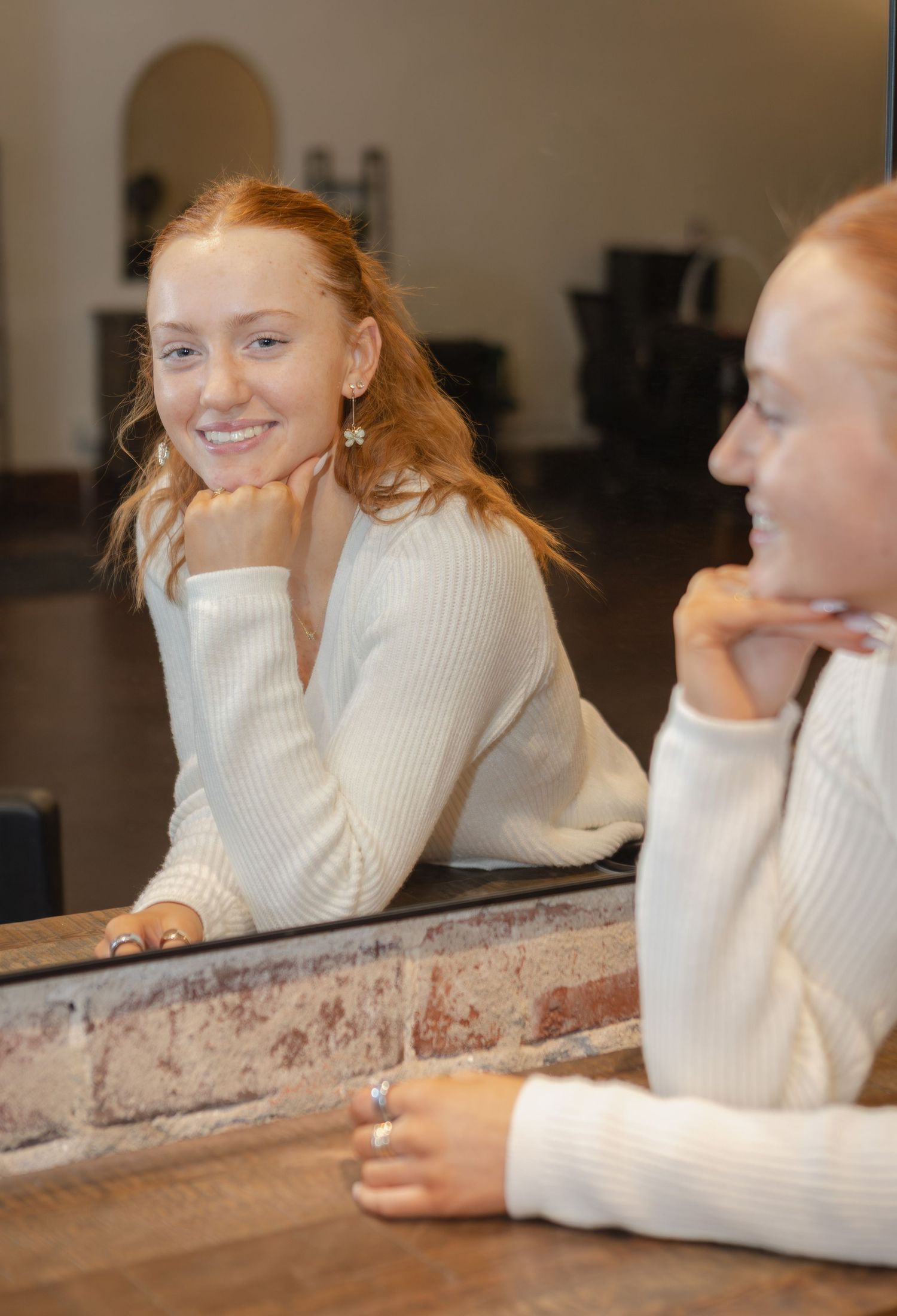 Redheaded young woman in white sweater smiling and leaning on counter with mirror reflection beside her.