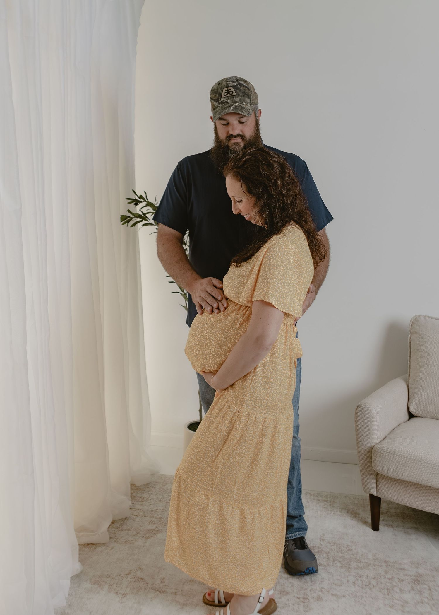 Couple sharing tender moment as man looks at pregnant woman in yellow dress near white curtains.