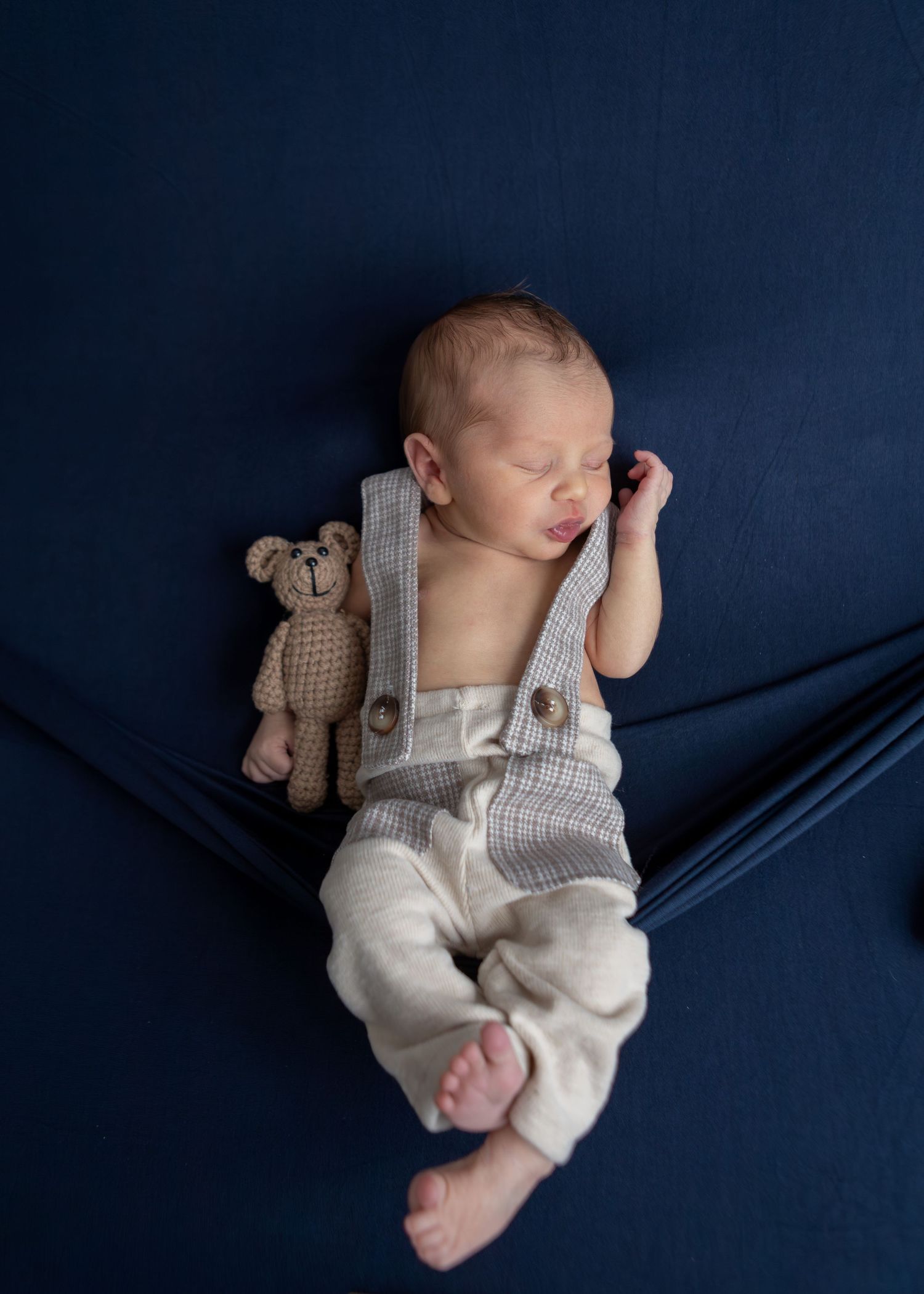 Sleeping newborn in suspender outfit lies beside knit teddy bear on navy blue fabric backdrop.