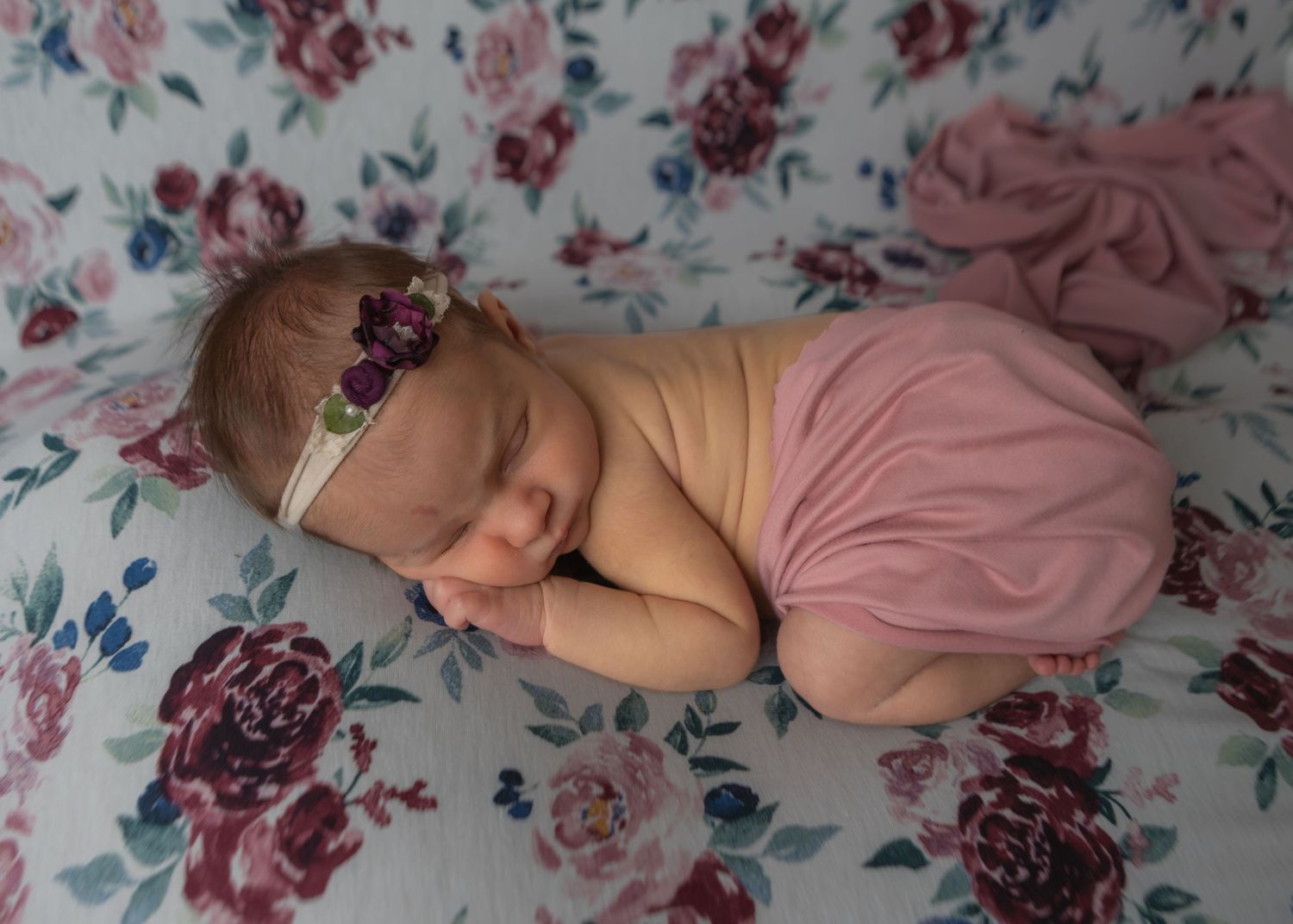 Sleeping newborn baby girl in pink wrap with floral headband on floral background.