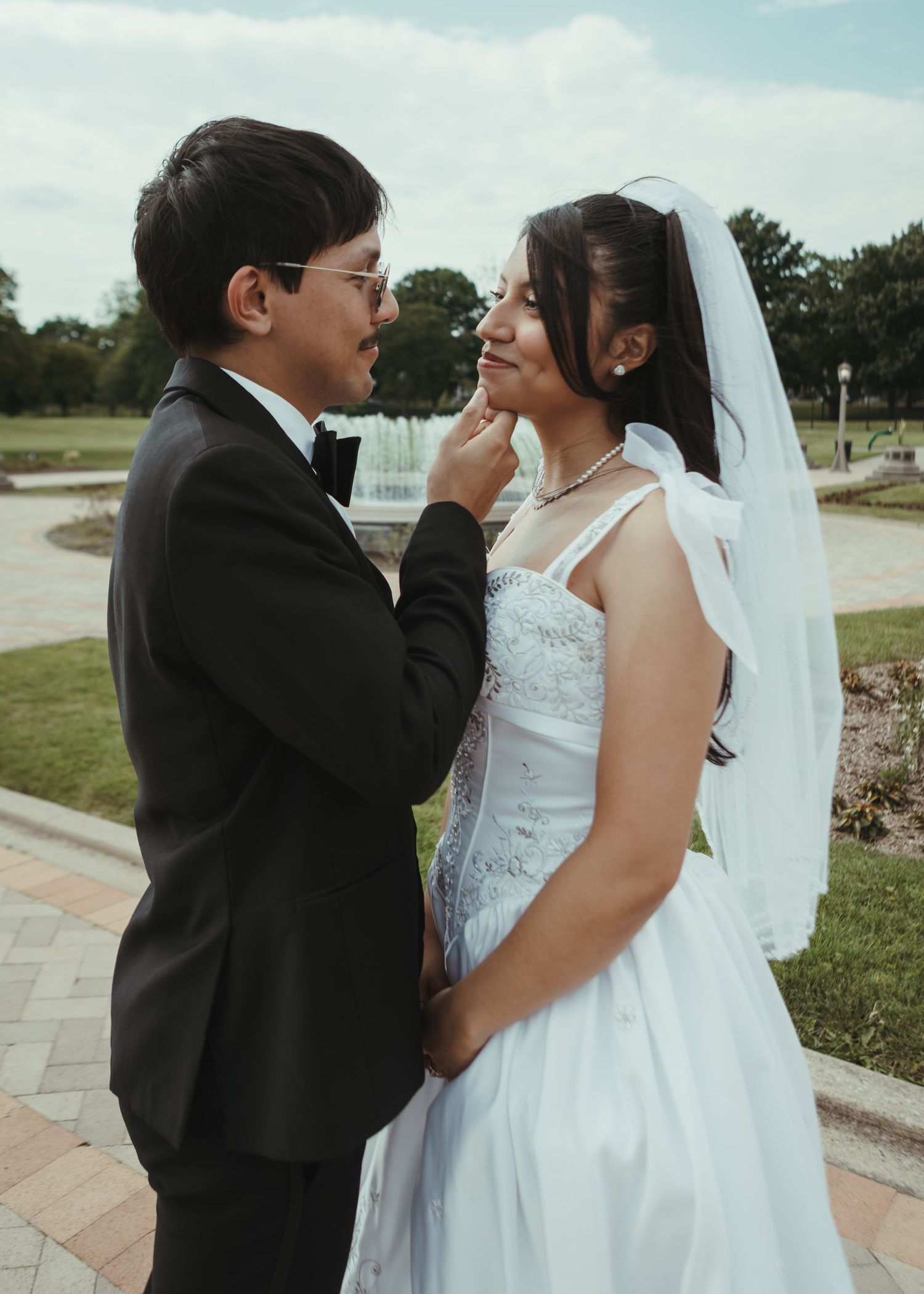 Newlywed couple in wedding attire share tender moment outdoors near fountain, groom touching brides face.