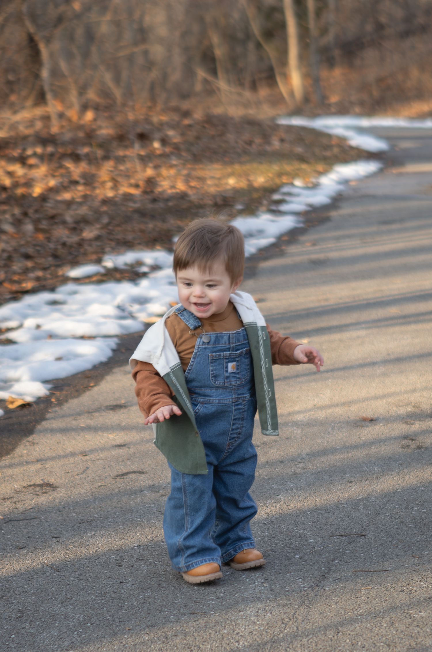 Smiling toddler in denim overalls walking on a path with patches of snow in background.