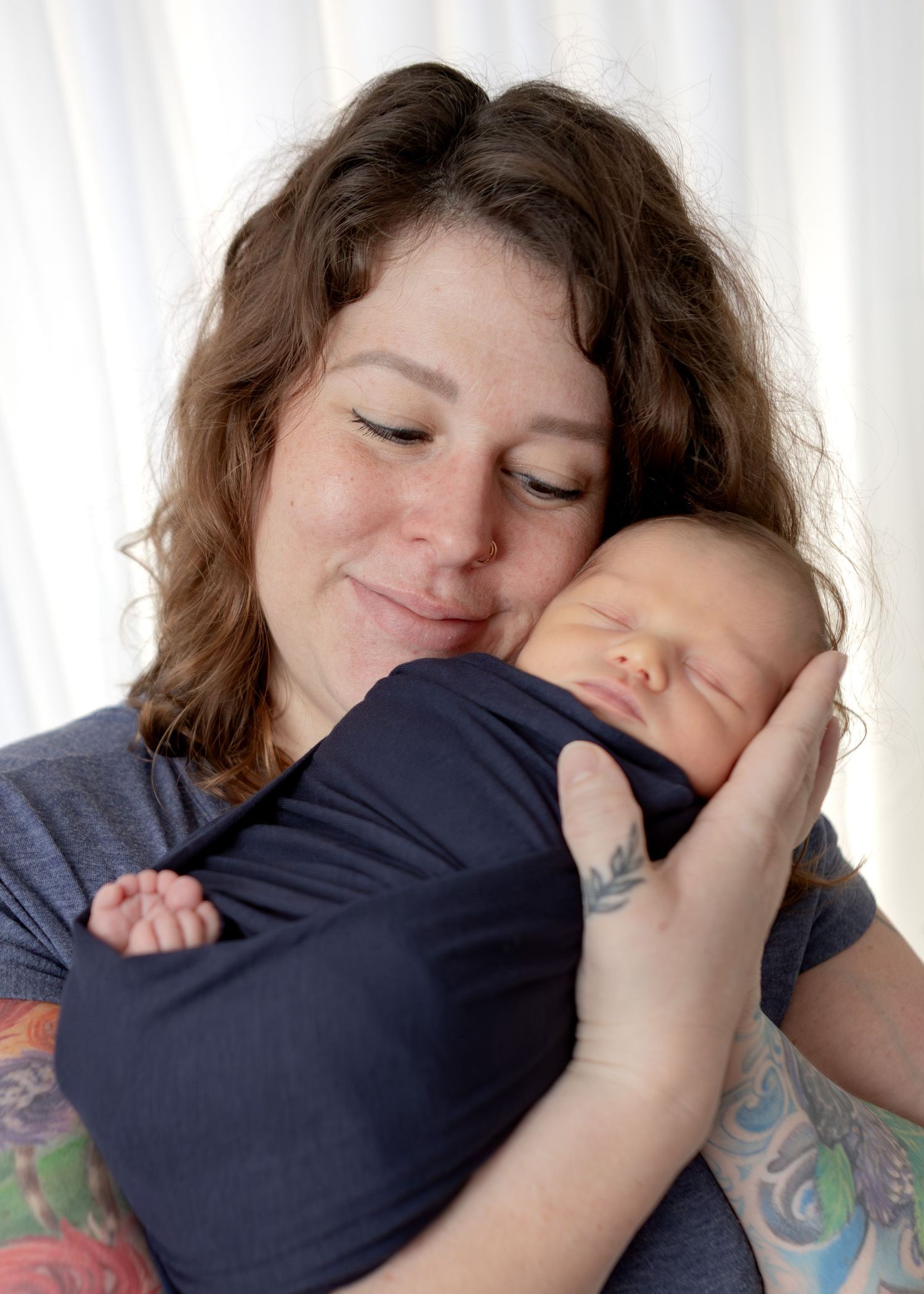 Loving mother with tattoos gently cradling her sleeping newborn baby wrapped in navy cloth.
