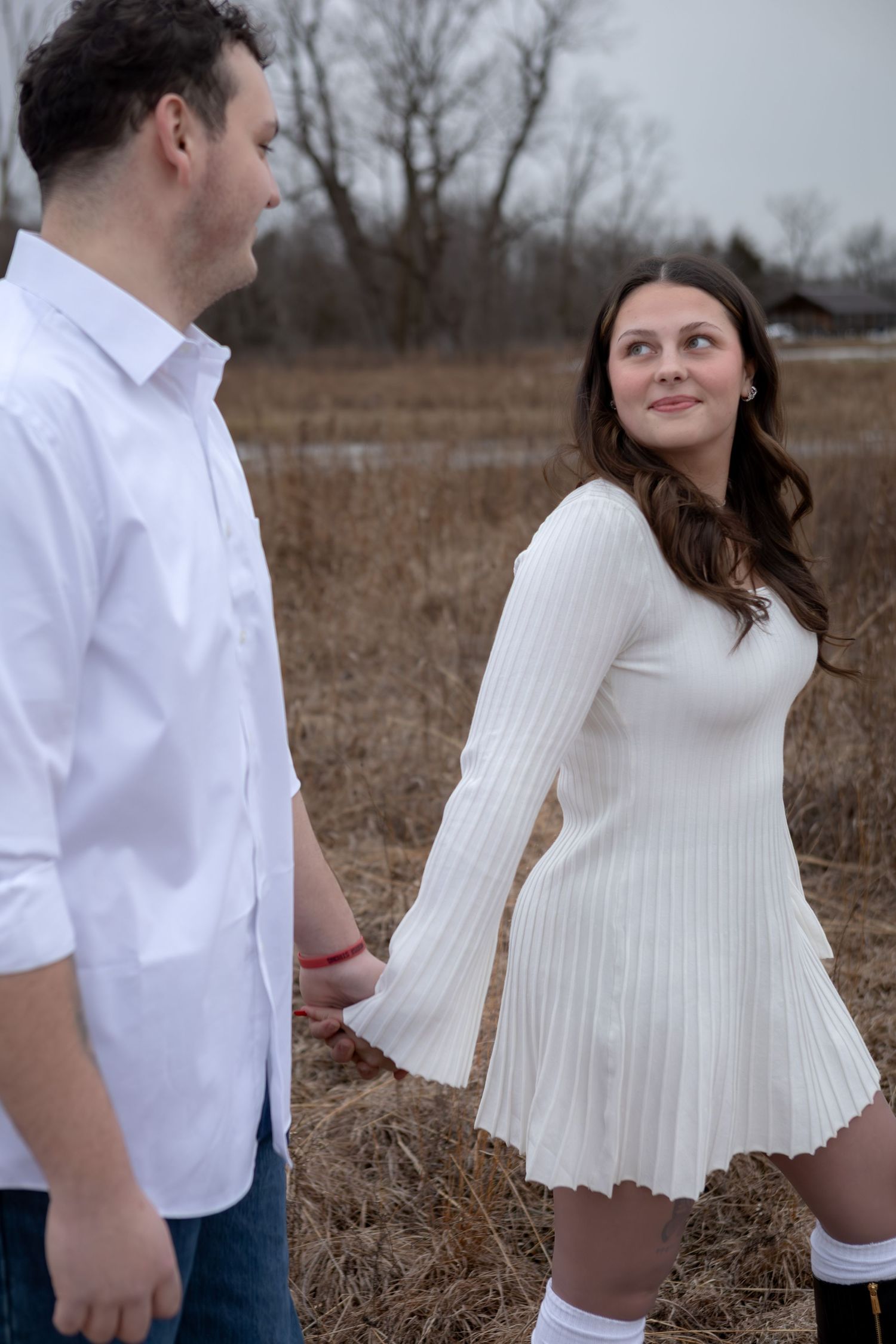 Couple holding hands in a winter field, woman wearing a white knit dress and knee-high socks.