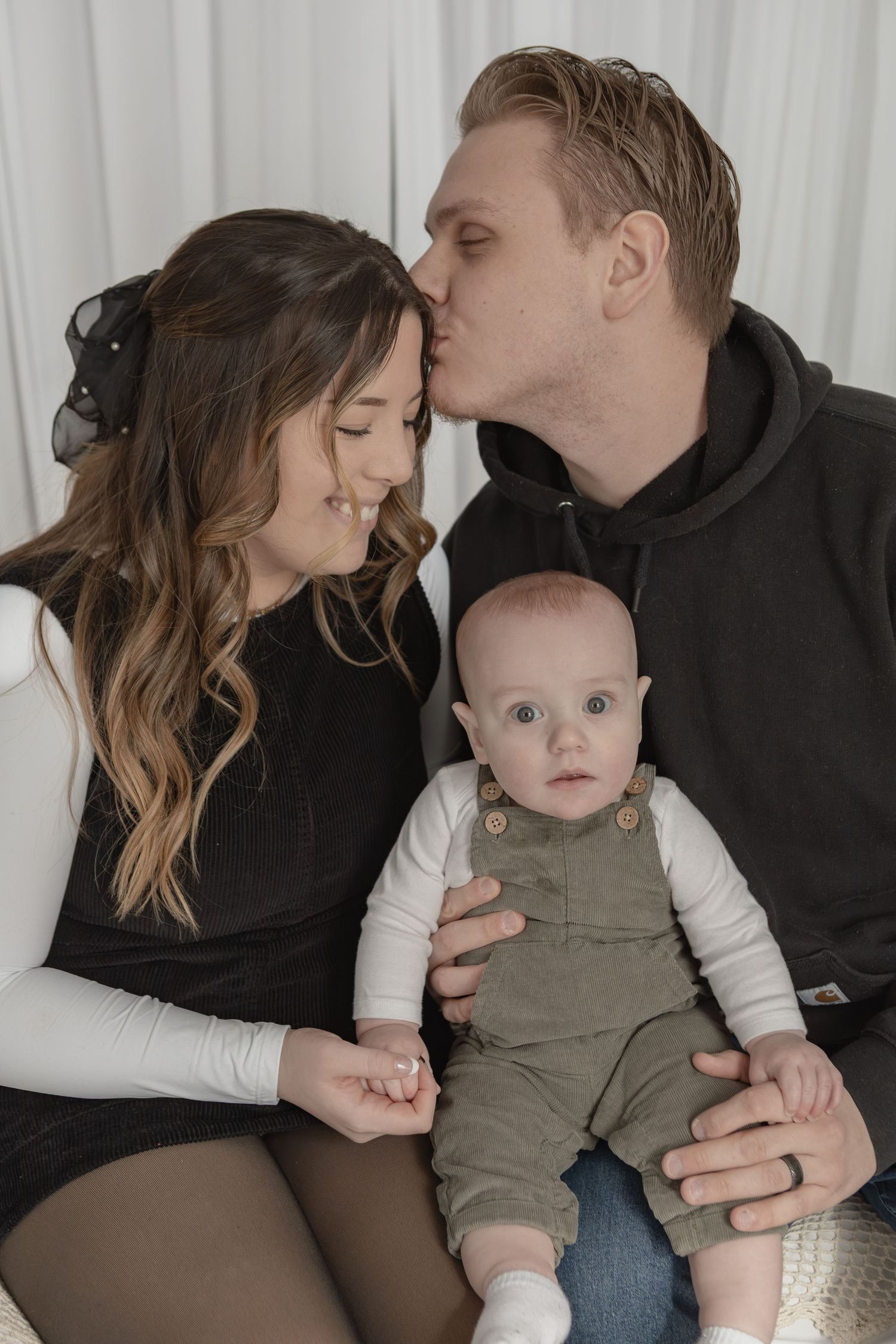 Parents kissing and holding their wide-eyed baby in olive overalls during a family portrait session.