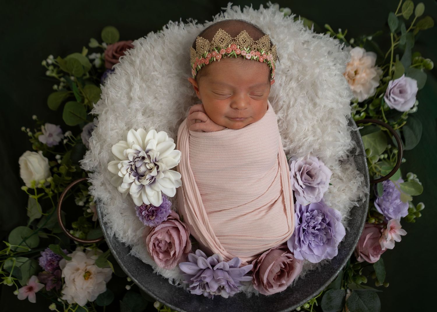 Newborn baby girl swaddled in pink wrap with floral crown, surrounded by roses in a white bowl.