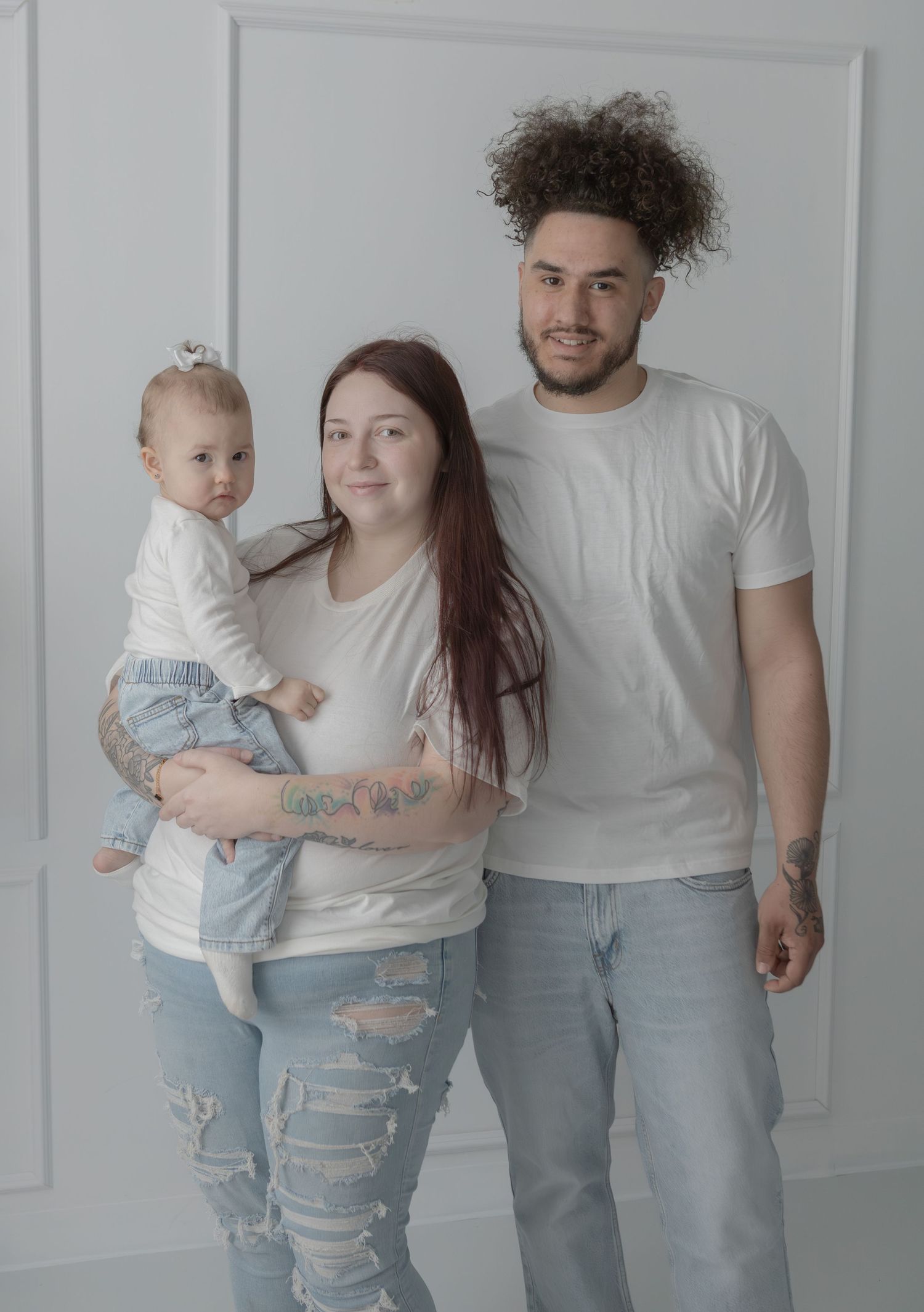 Young family of three in white tees and jeans posing together in a bright white studio.