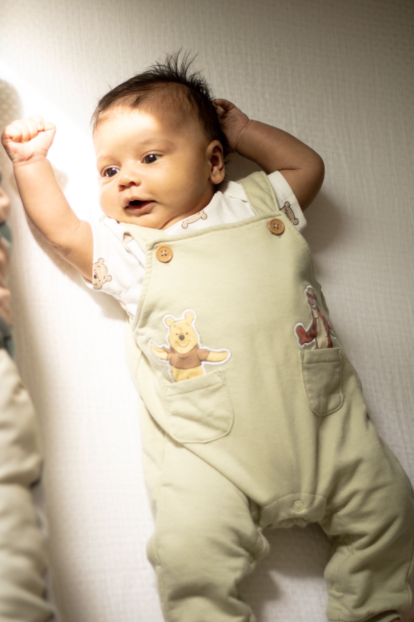 Happy newborn baby lying on white blanket wearing Winnie the Pooh green overalls with arm raised.