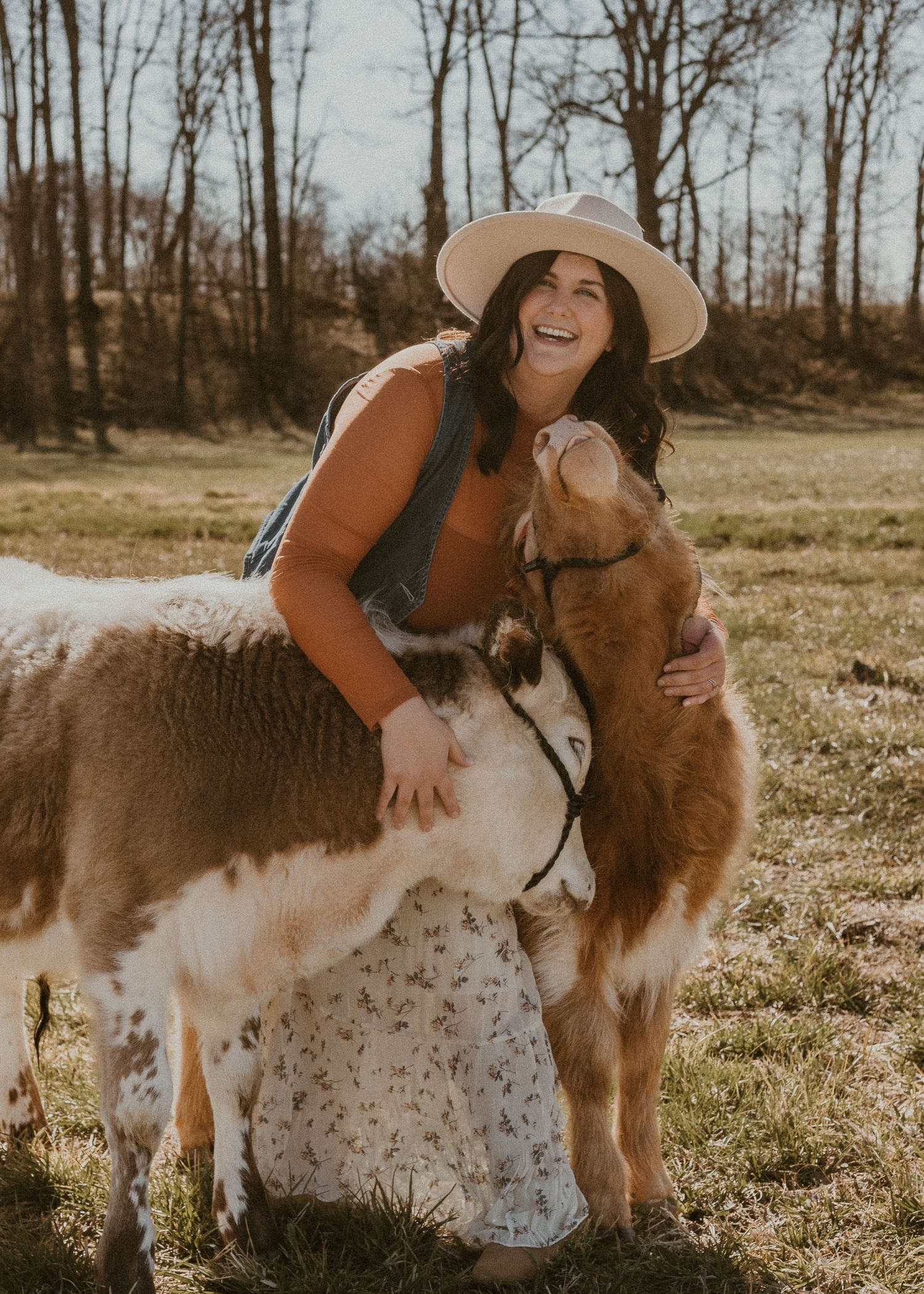 Laughing woman in hat hugging a miniature cow and dog in a sunny field.