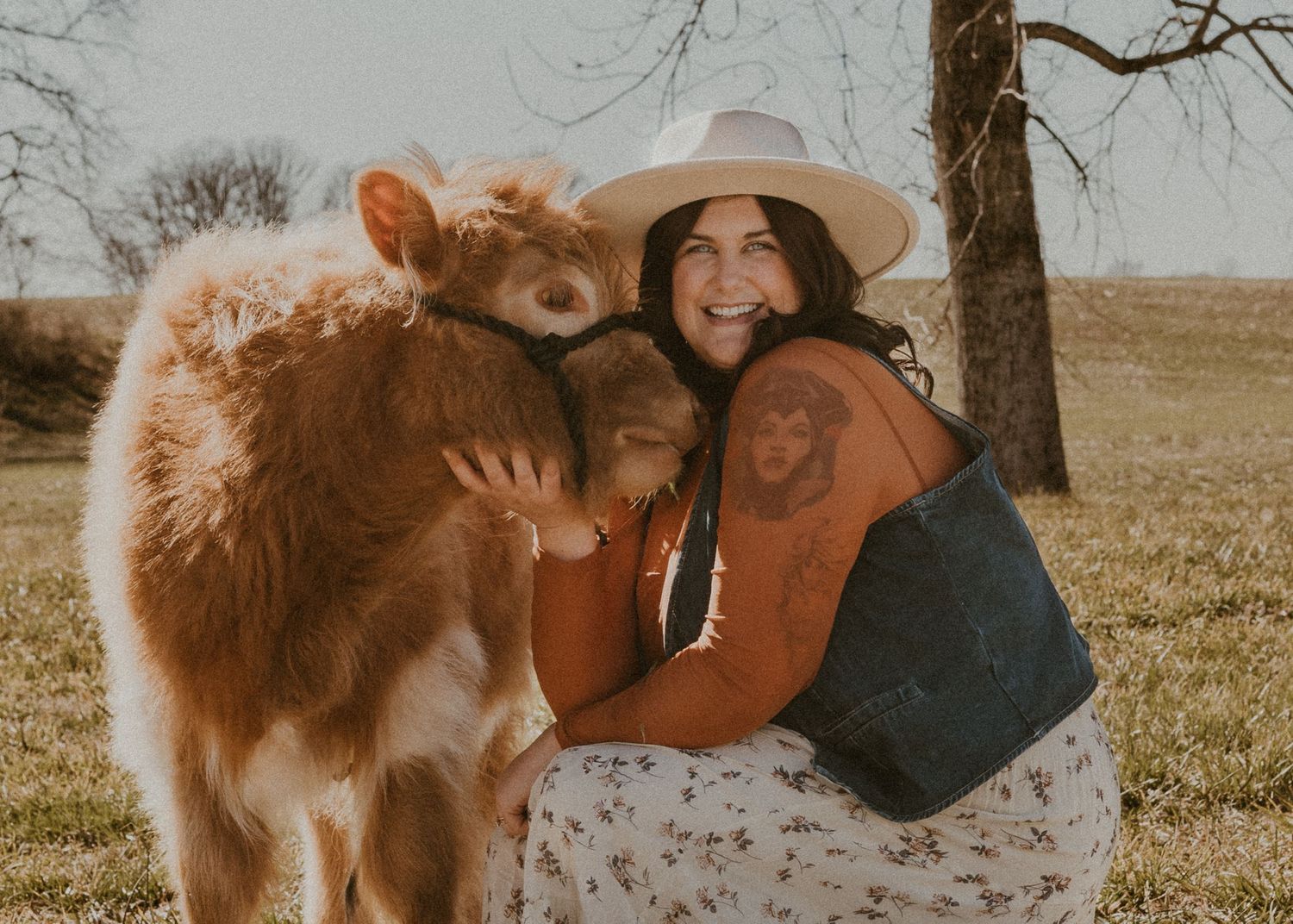 Smiling woman in a white hat hugging a fluffy brown Highland calf in an open field.