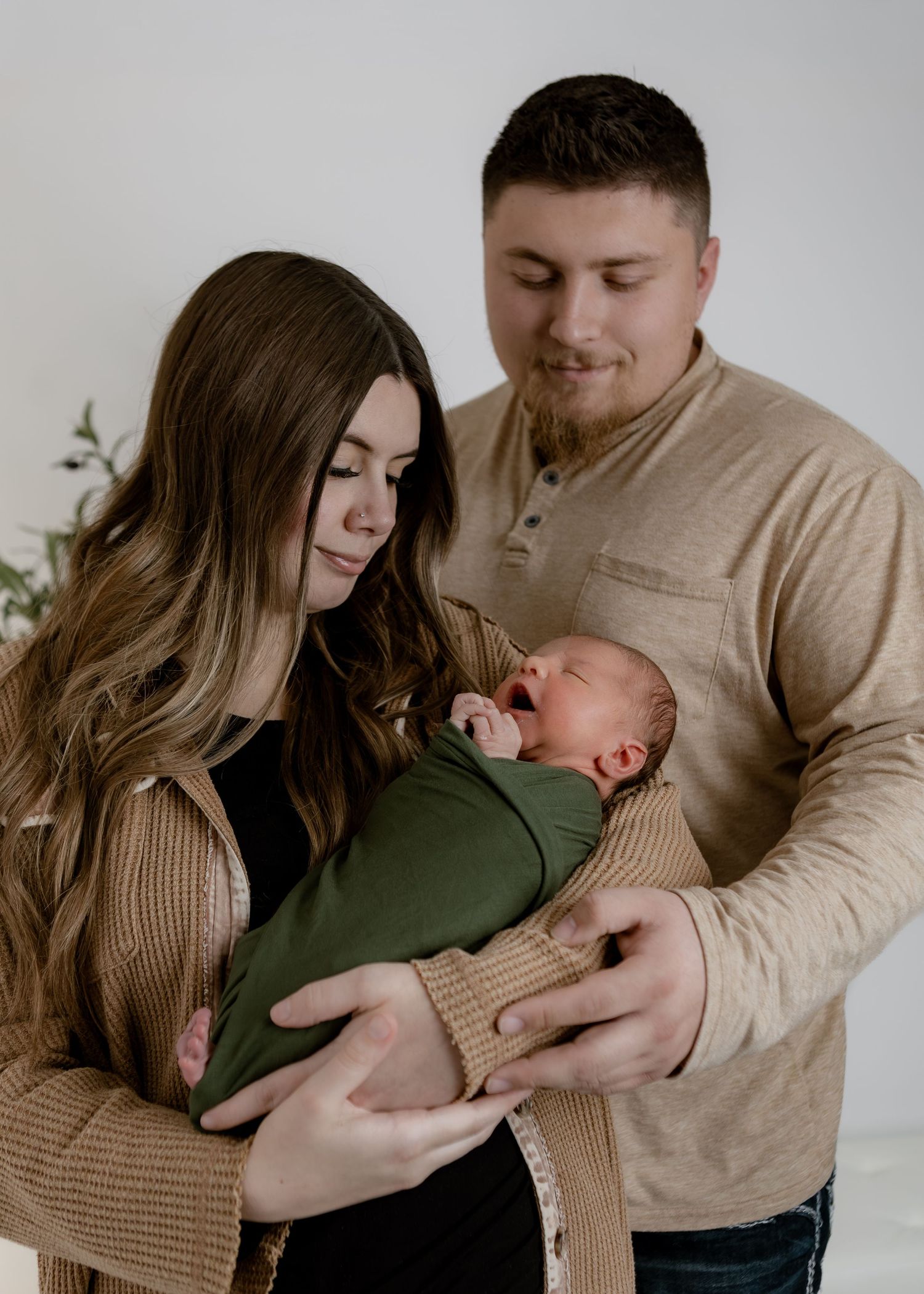 New parents in neutral tones lovingly gaze at their green-swaddled newborn baby indoors.