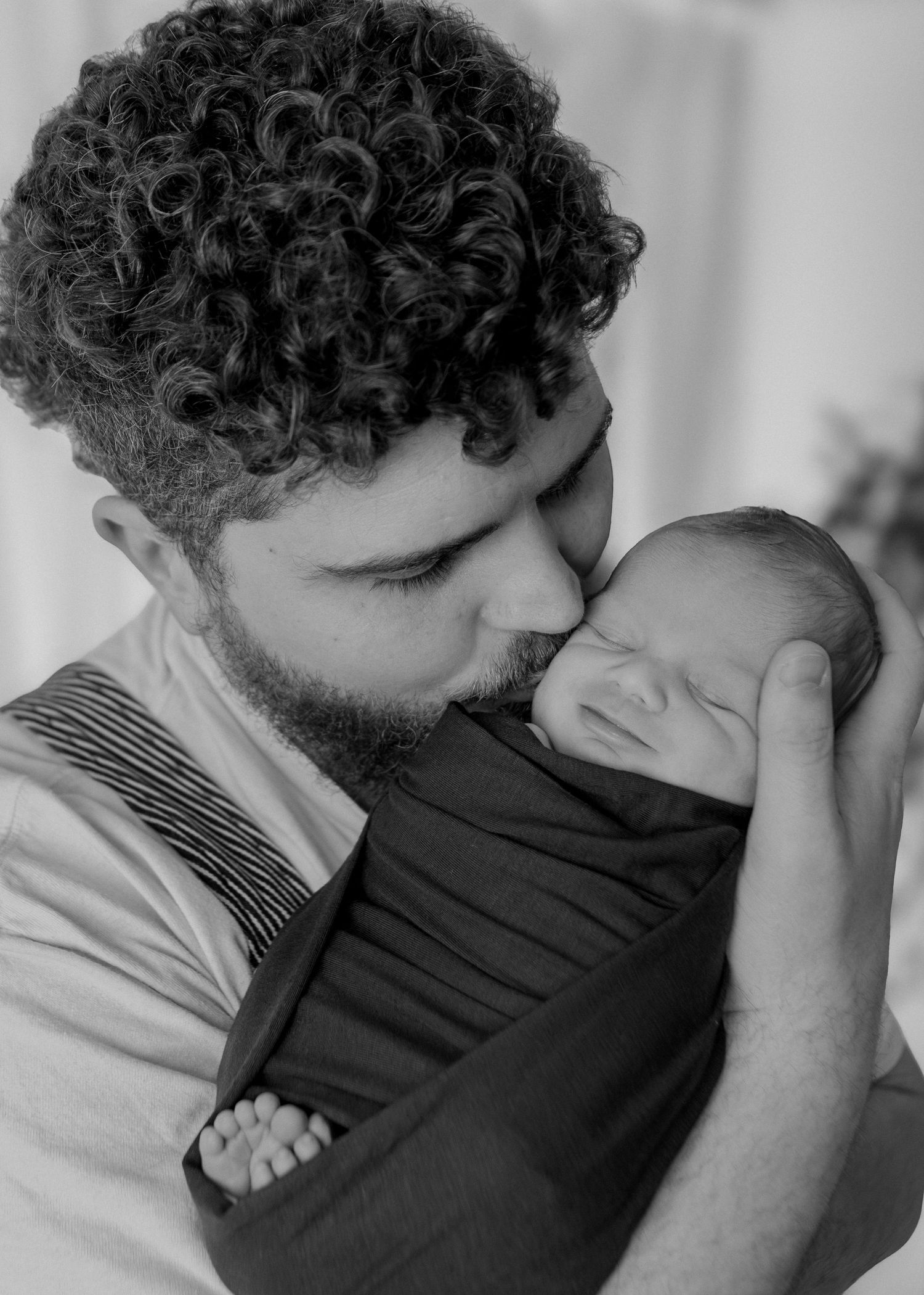 Black and white photo of a curly-haired father gently kissing his sleeping swaddled newborn.