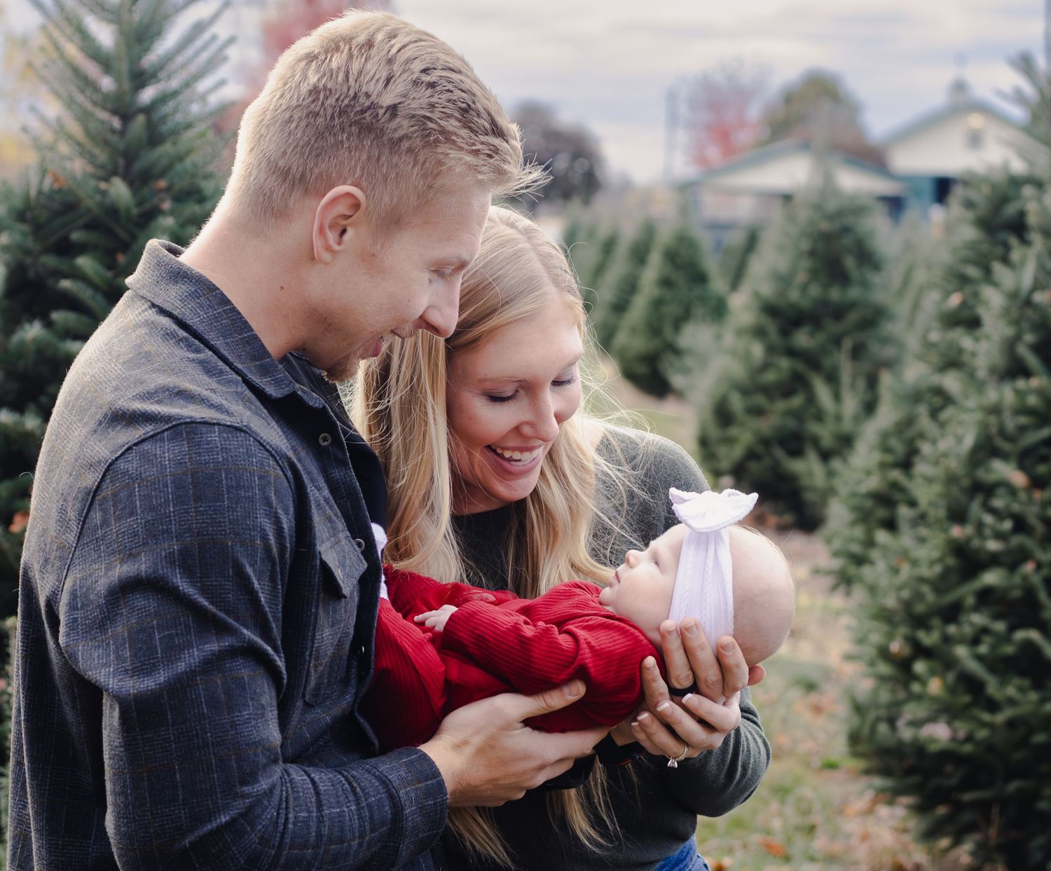 Young couple smiles at their newborn baby dressed in red at an outdoor Christmas tree farm.