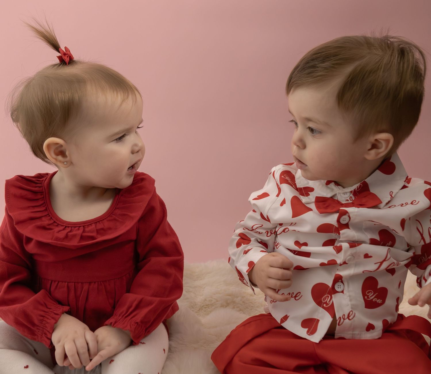 Two adorable babies in red Valentine's Day outfits sitting together on a pink background.