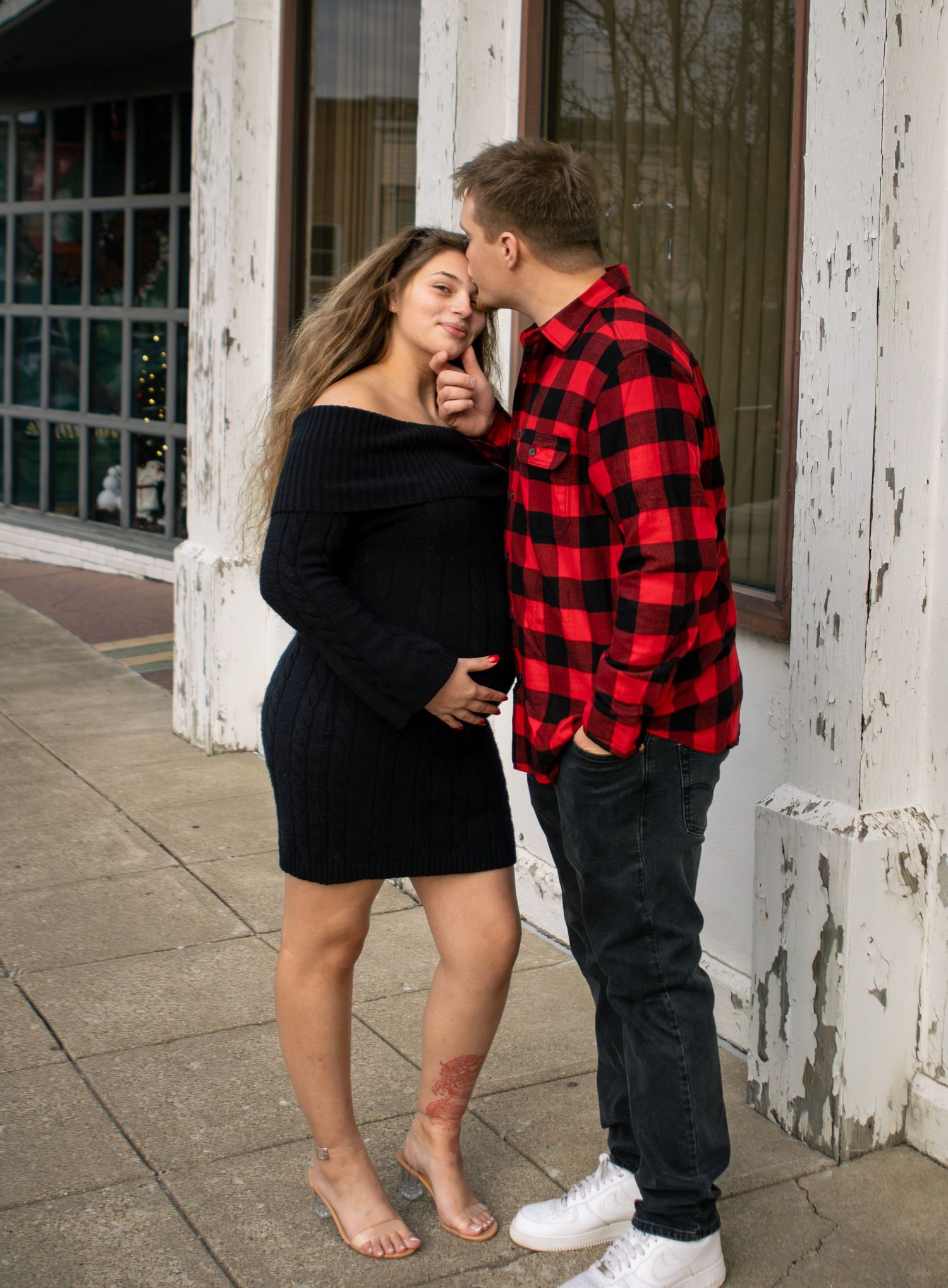 Couple sharing a tender moment on a sidewalk, man in plaid shirt kissing pregnant woman in black dress.