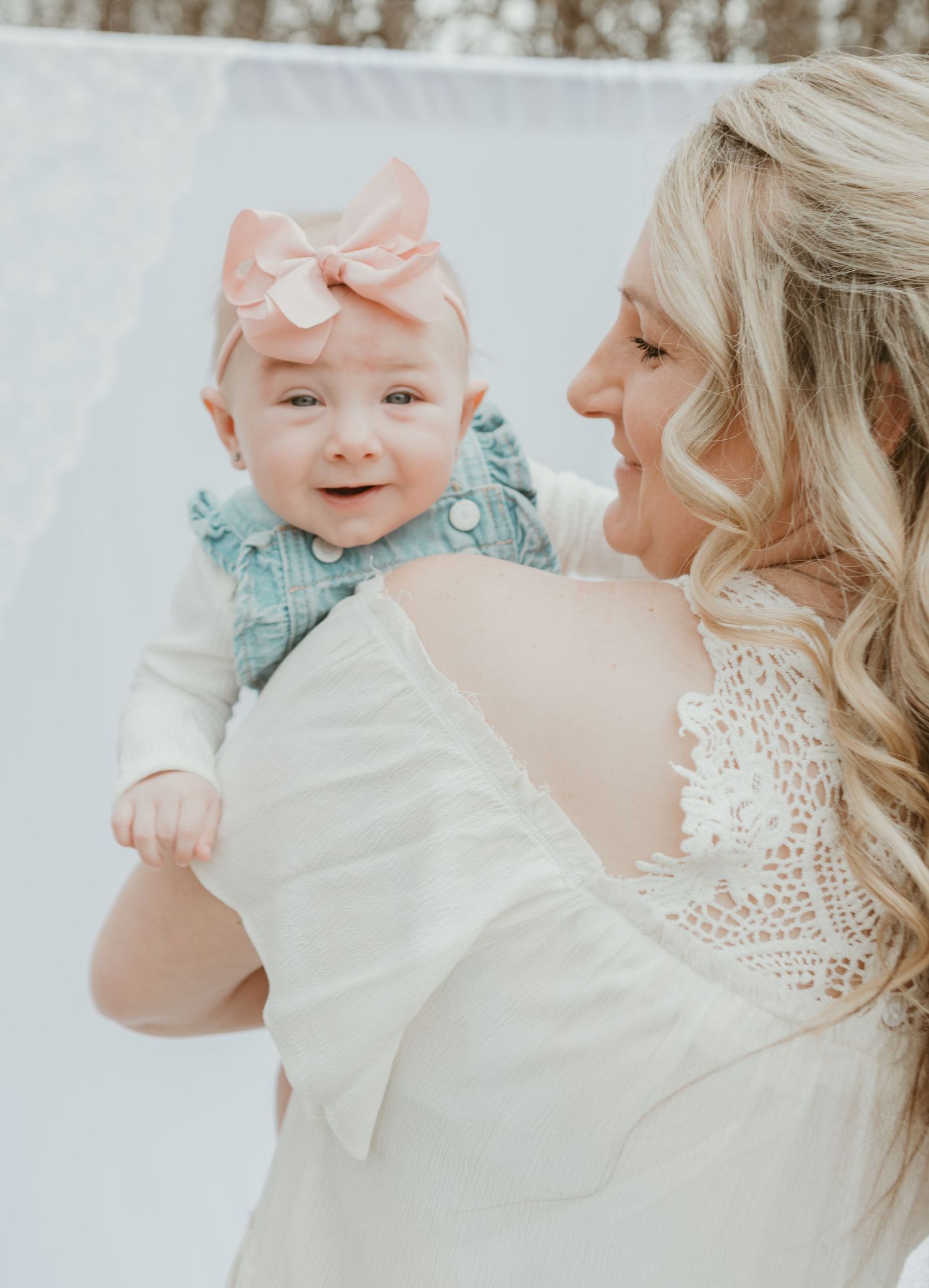 Smiling blonde mother in white lace dress holding happy baby girl with pink bow.