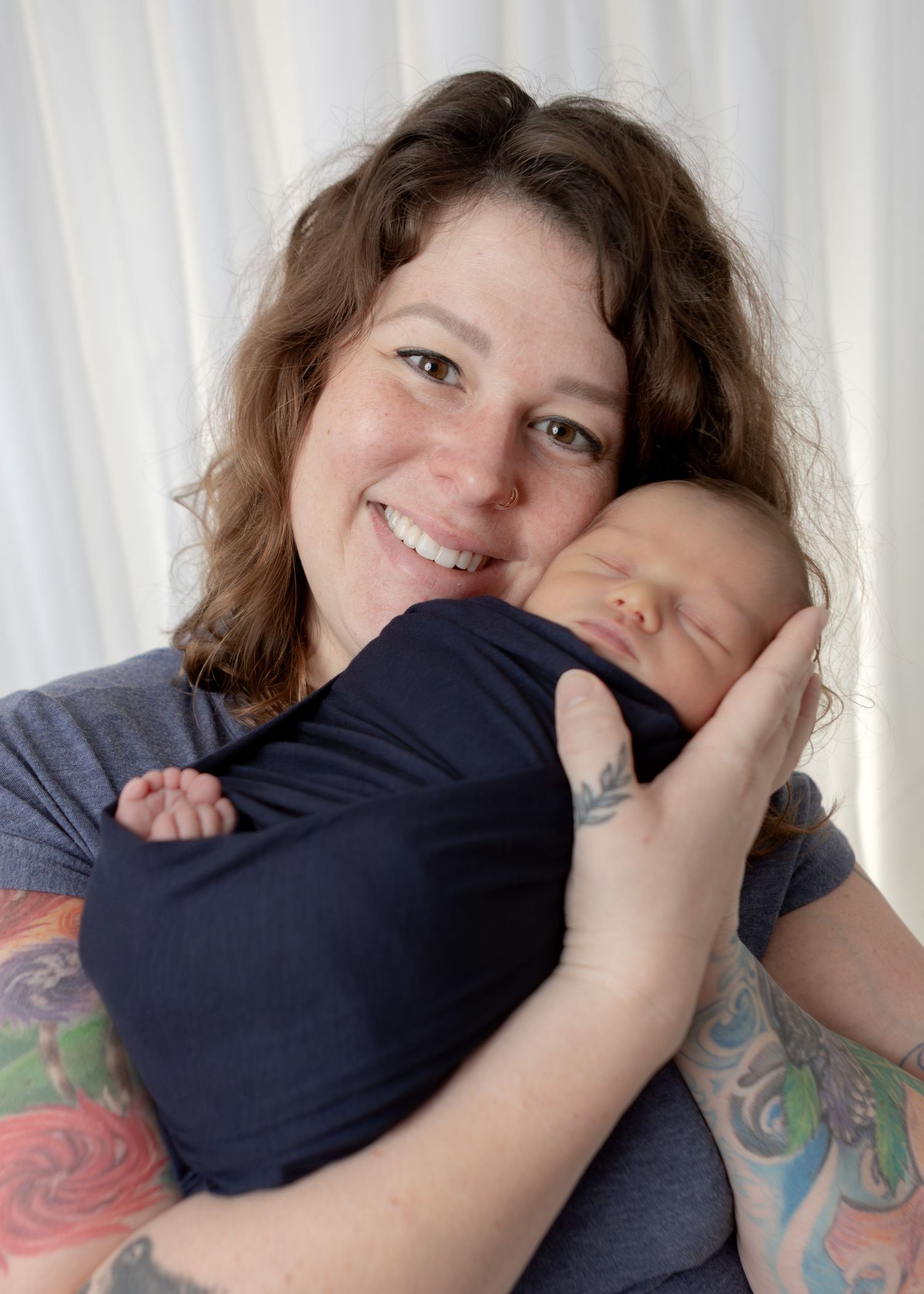 Smiling tattooed mother holds sleeping newborn swaddled in navy blue wrap close to her cheek.