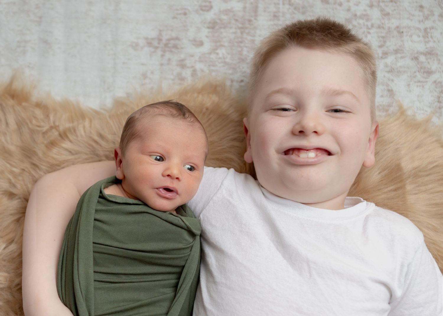 Smiling young boy lying beside his newborn sibling wrapped in a green swaddle on a fur rug.