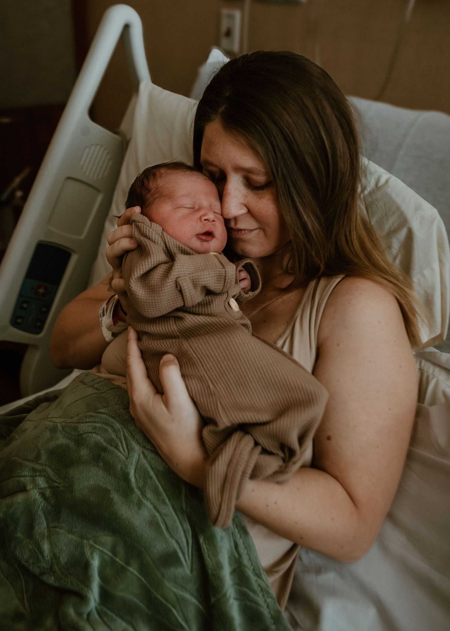 New mother in hospital bed tenderly holds and nuzzles sleeping newborn baby wrapped in brown.