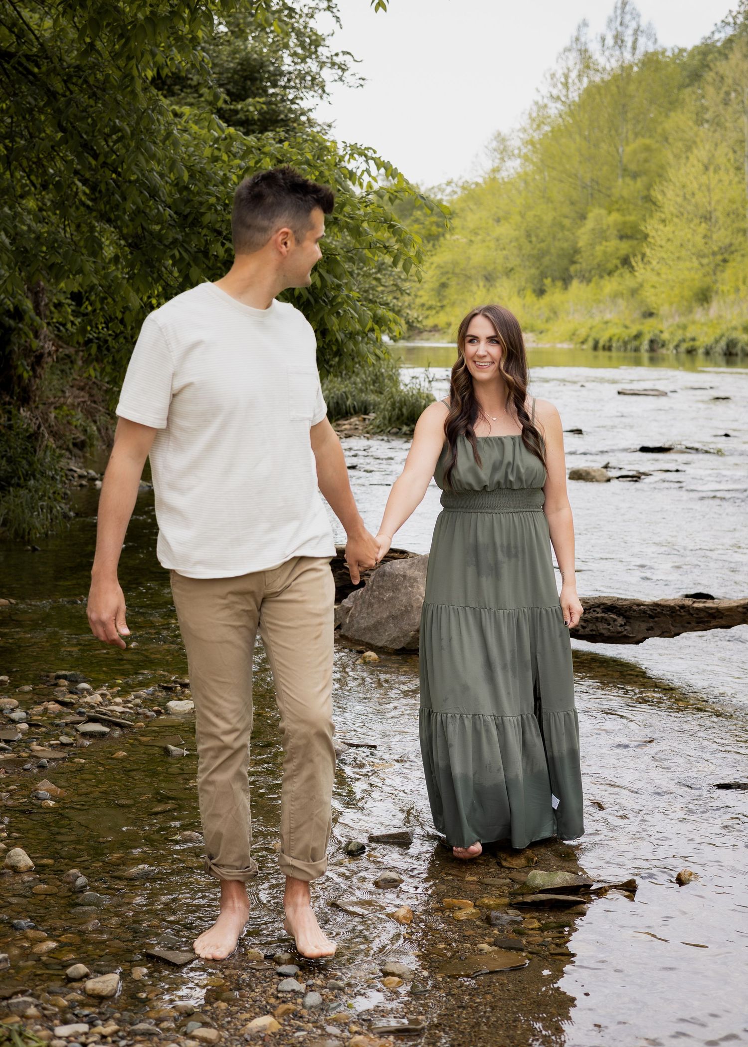 A couple holds hands while walking barefoot along a scenic rocky riverbank with green trees.