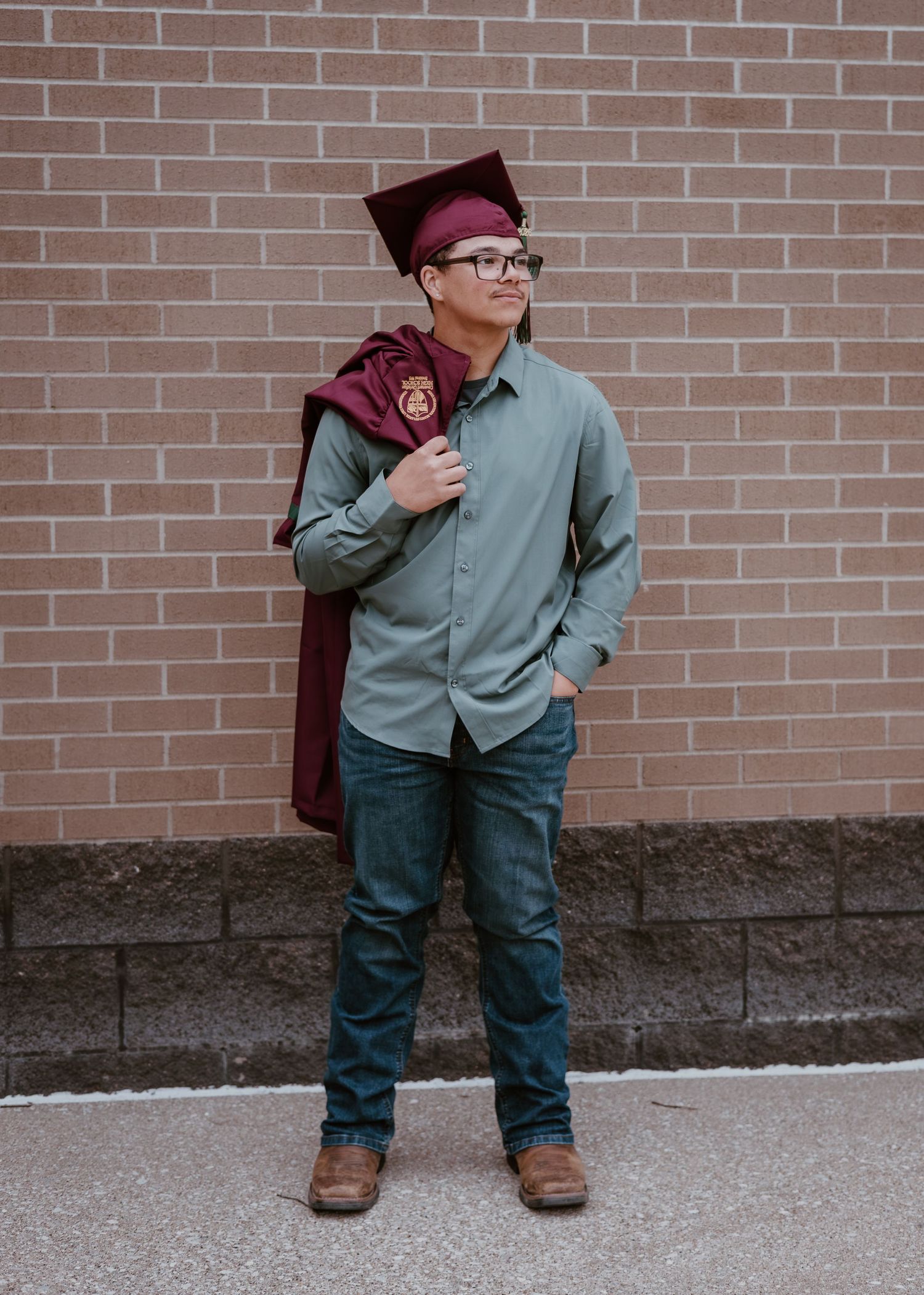 A young man in glasses holds his maroon graduation gown over his shoulder against a brick wall.