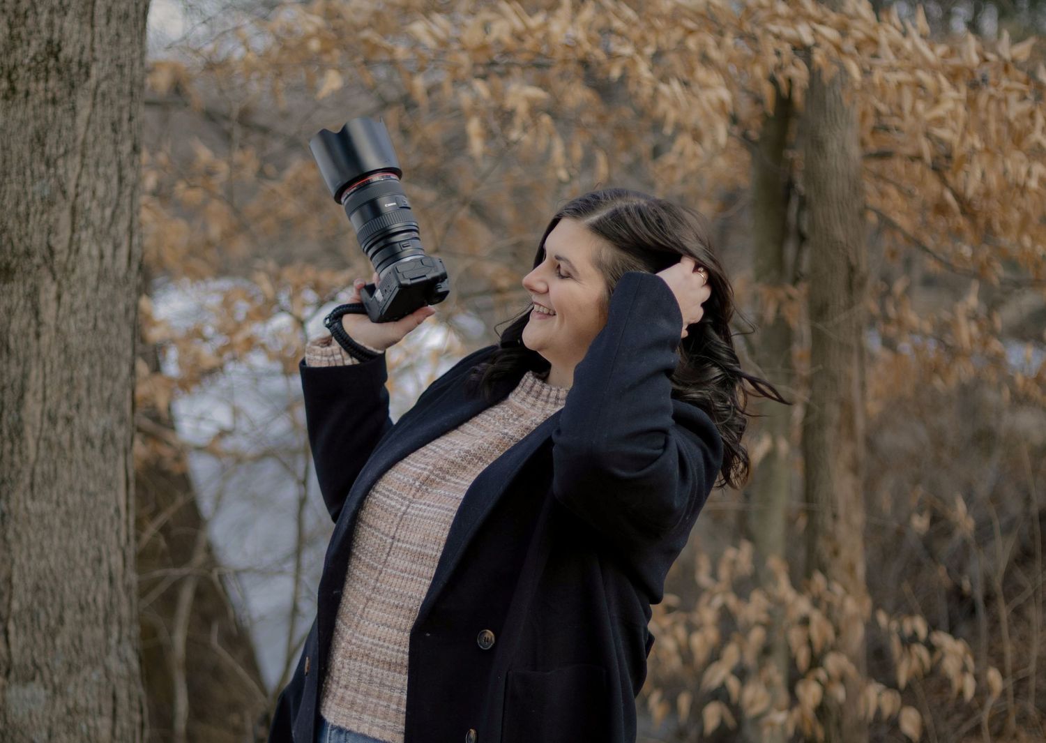 Female photographer holding camera up outdoors among autumn trees with wind blowing her hair.