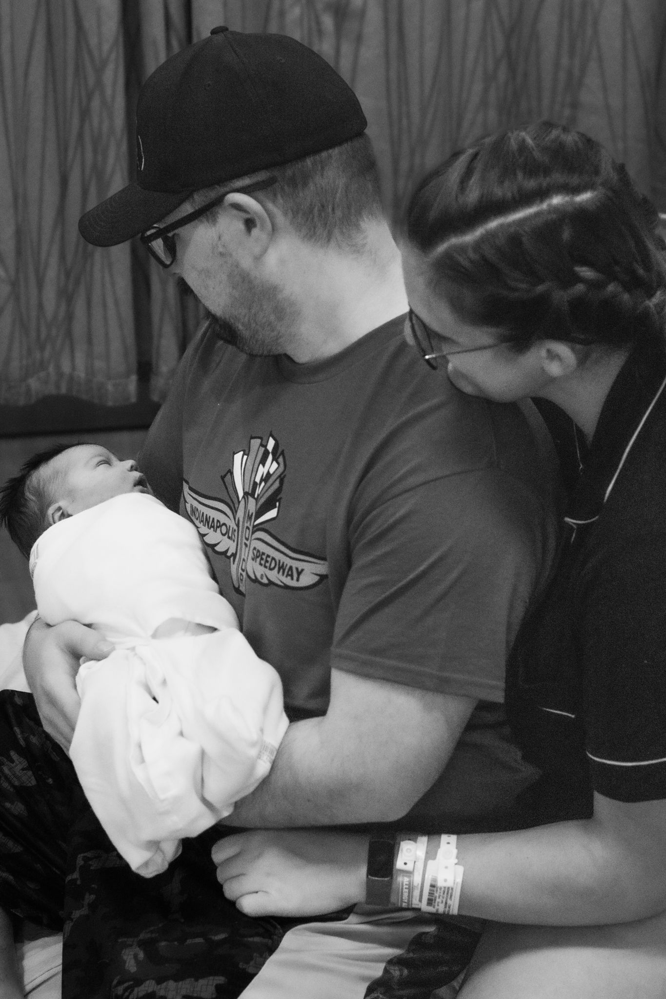 Black and white photo of new parents lovingly gazing at their swaddled newborn baby in a hospital room.