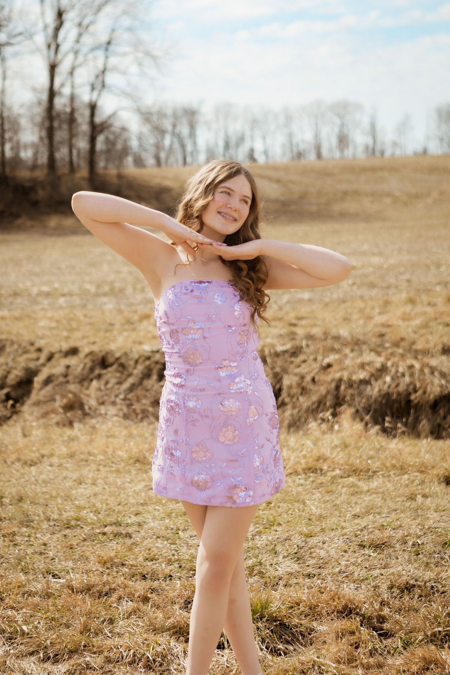 Smiling young woman in a purple floral mini dress poses with hands behind her neck in an open field.
