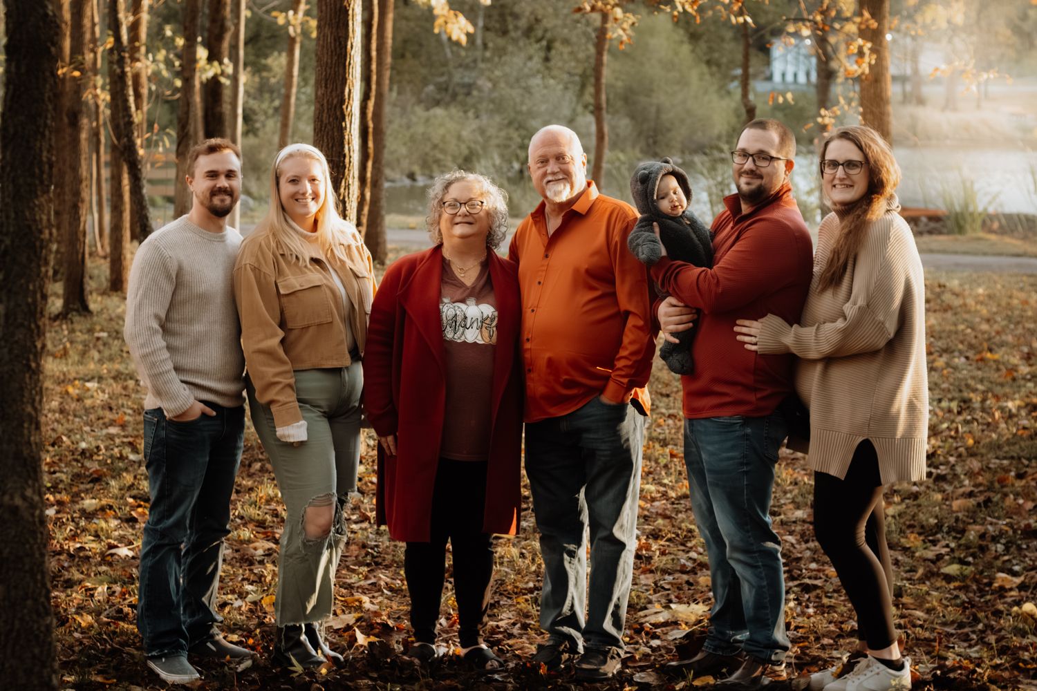 Multi-generational family of six smiling together outdoors among autumn foliage in wooded setting.