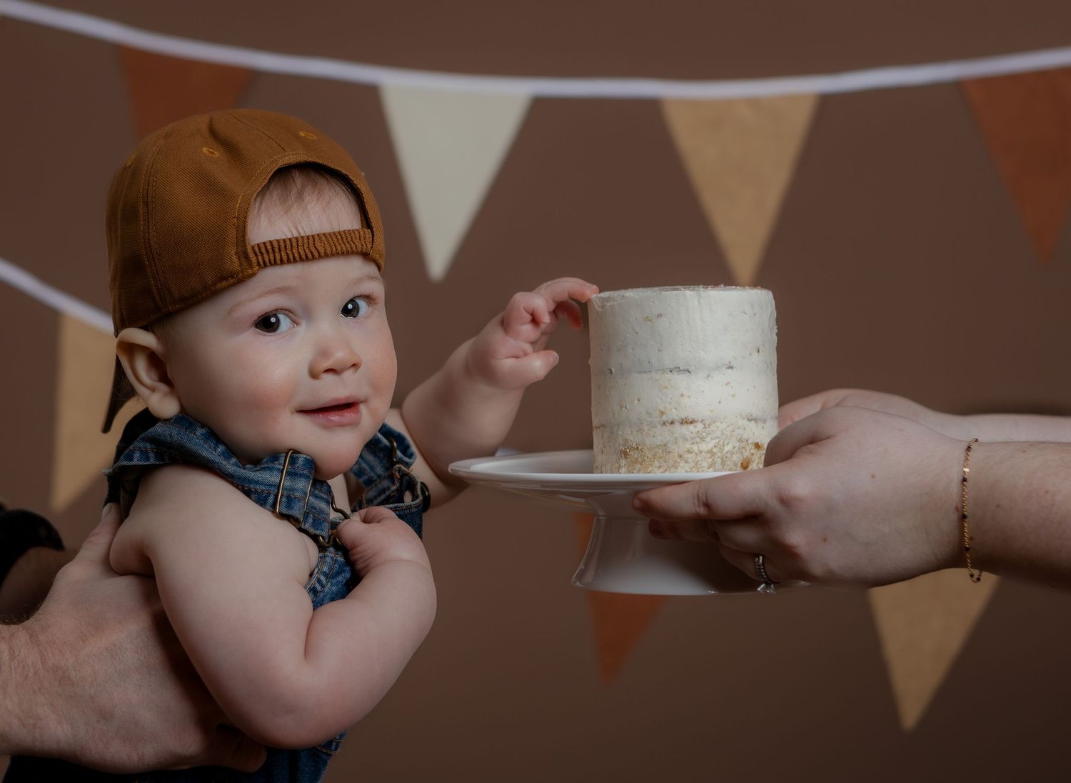 Curious baby boy in denim overalls and brown cap reaching for a white birthday cake being presented to him.