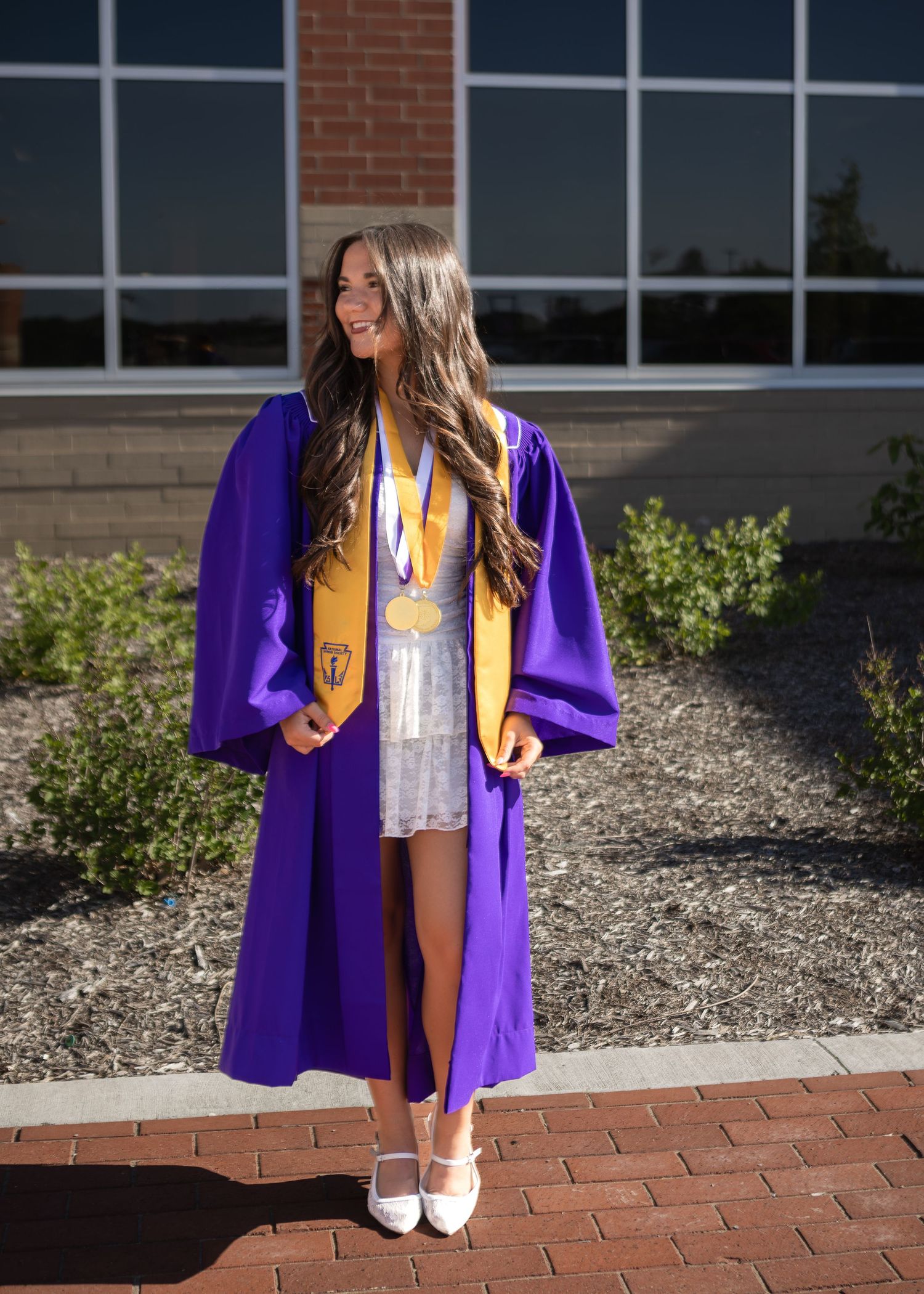 Female graduate in full purple cap and gown with gold stole and medals stands outside sunny building.