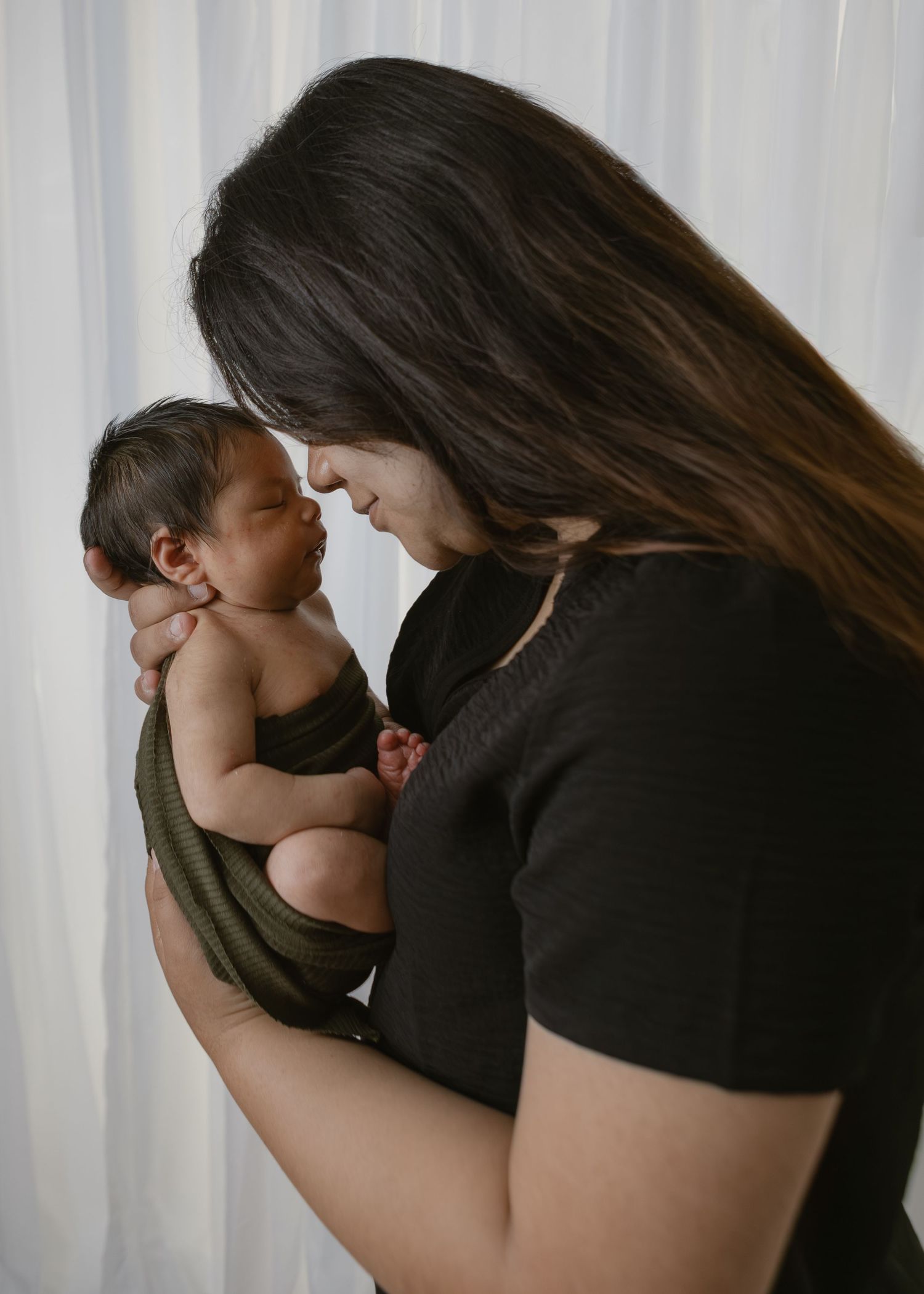 Mother tenderly holding her newborn baby wrapped in a green swaddle near sheer curtains.