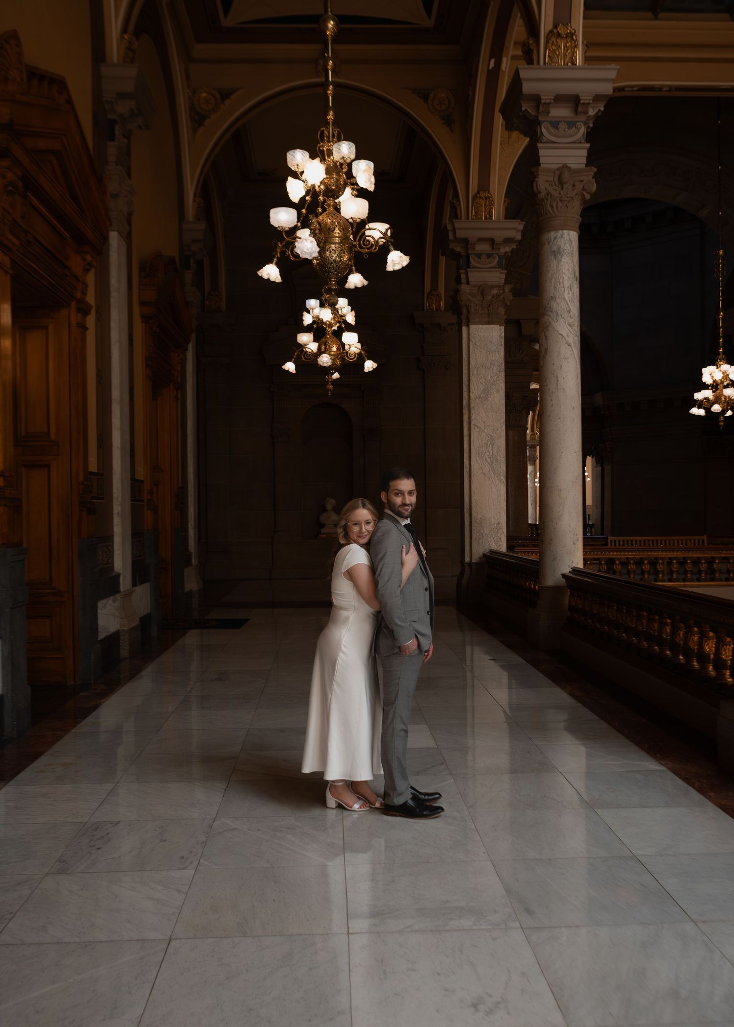 Bride and groom standing back-to-back in an elegant chandelier-lit marble hallway of a grand historic building.