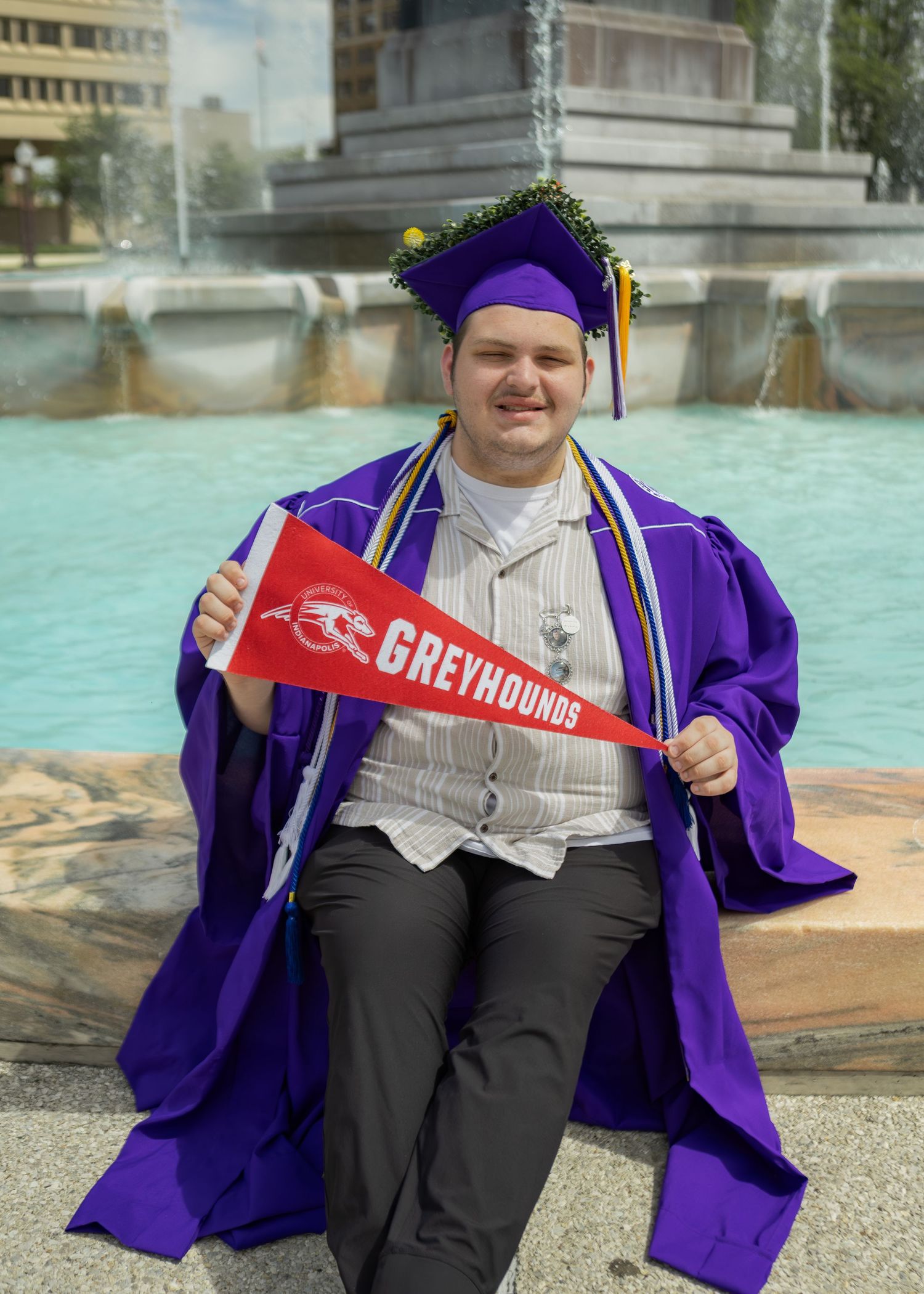 Male graduate in purple cap and gown sits by fountain holding red Greyhounds pennant flag.