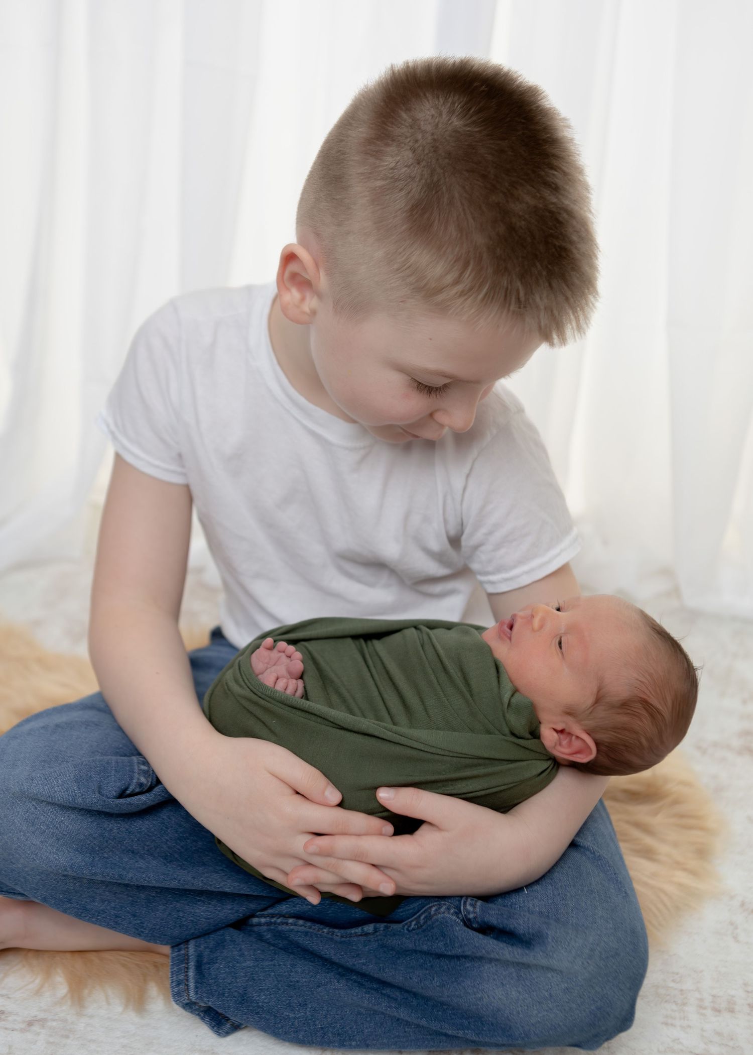 Young boy in white tee sits cross-legged holding sleeping newborn swaddled in green wrap.