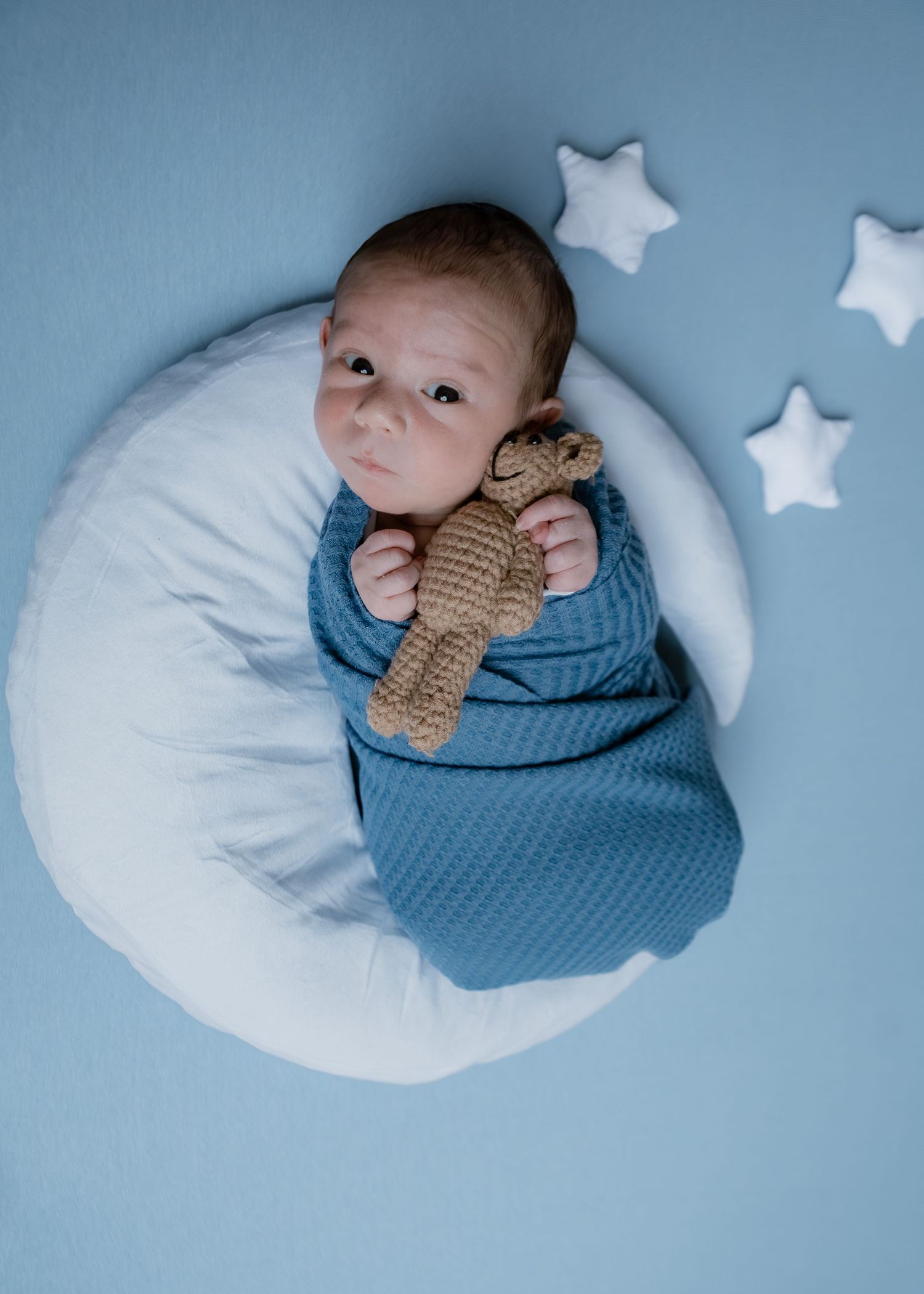 Newborn baby swaddled in blue on moon pillow with teddy bear and star props.