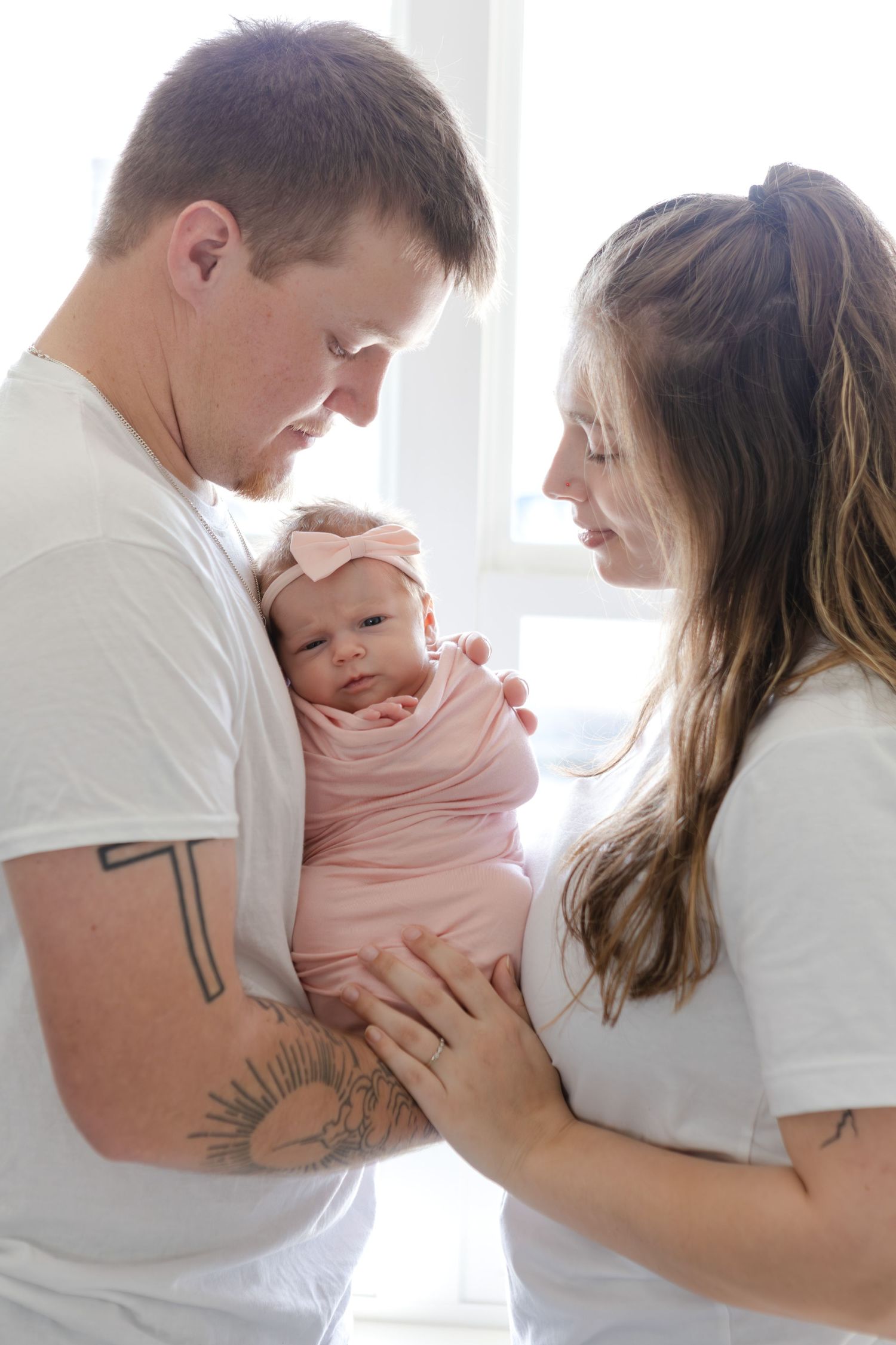 Two parents in white shirts gently hold their pink-swaddled newborn baby near a bright window.