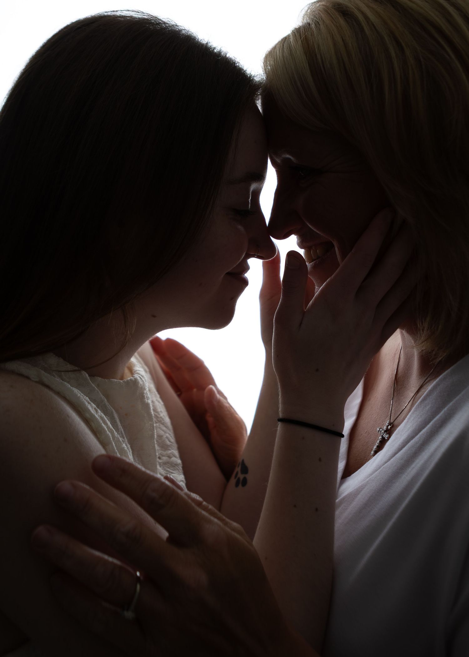 Two women smiling with foreheads touching in a tender backlit silhouette portrait.