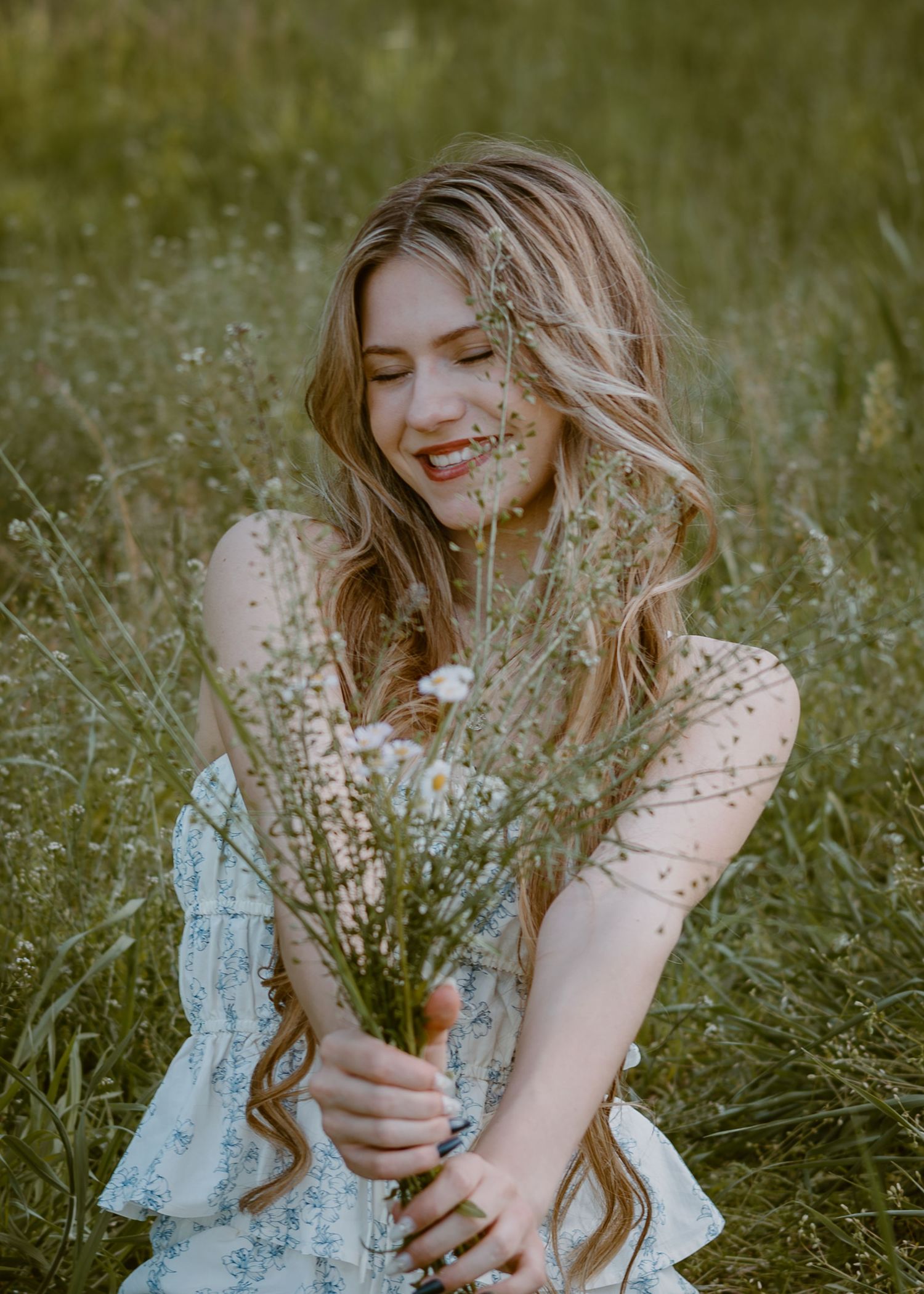 Smiling young woman with wavy blonde hair holding wildflowers while sitting in a lush green meadow.