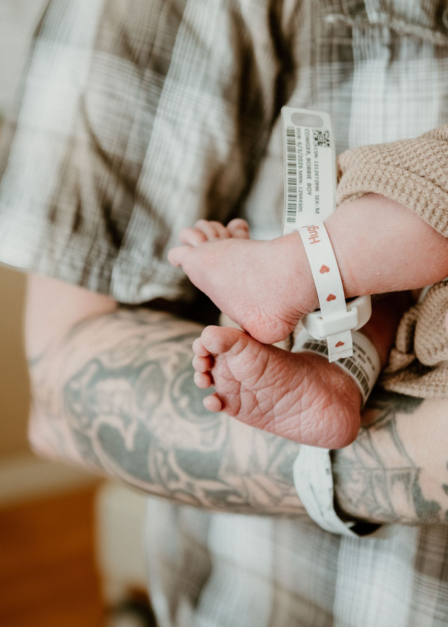 Tattooed father cradles tiny newborn feet with hospital ID bracelet on his forearm.