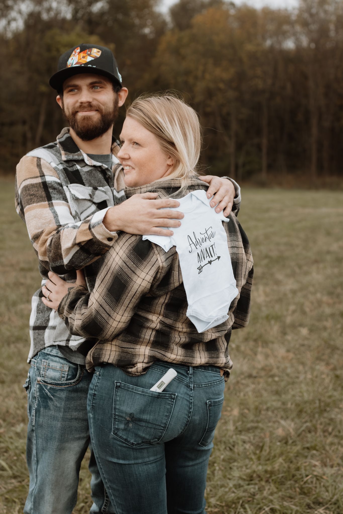Couple in matching flannel shirts embracing in a field, holding a baby onesie announcement.