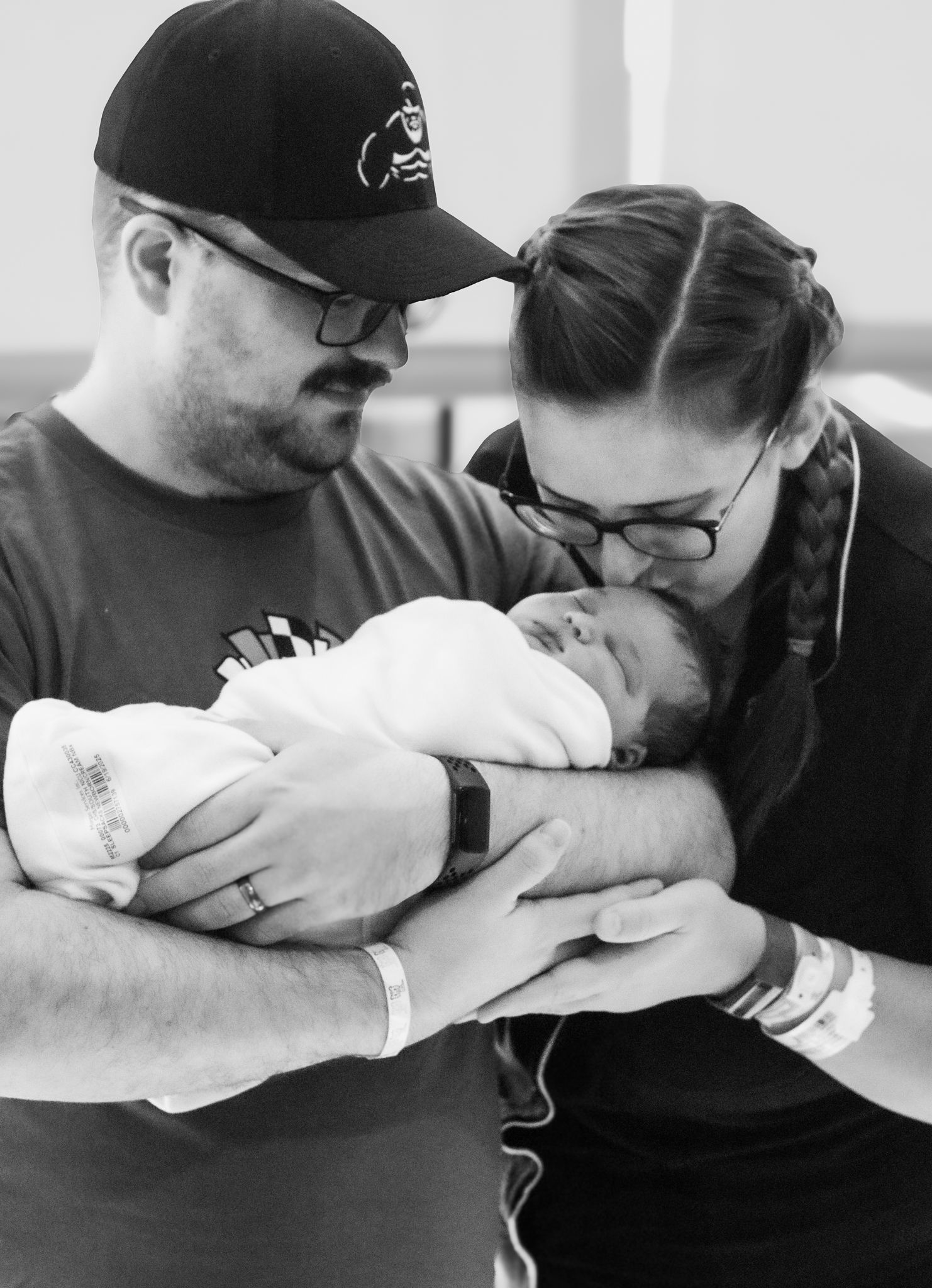 Black and white photo of new parents tenderly holding their swaddled newborn baby in a hospital room.