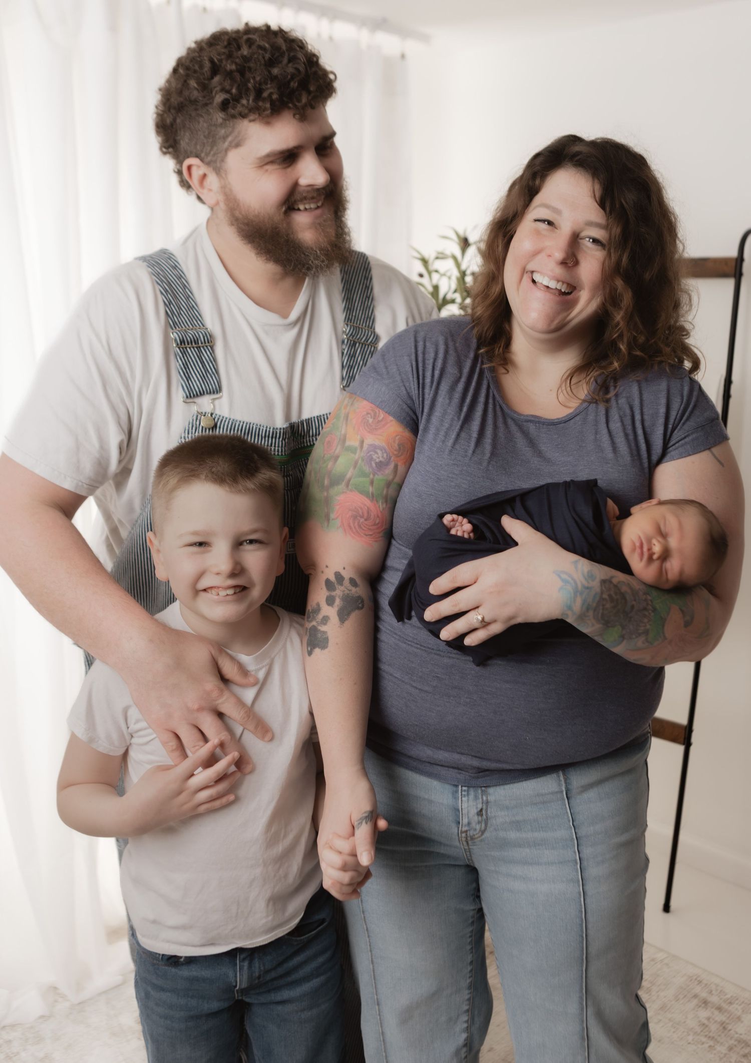 Tattooed couple smiling with young son and newborn baby in a bright indoor photo session.
